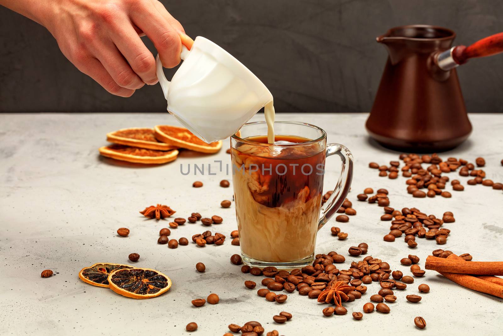 In a freshly made espresso in a transparent glass cup, a man's hand adds white cream, surrounded by coffee beans, dried slices of orange and lemon, anise stars and cinnamon sticks on a gray concrete background.