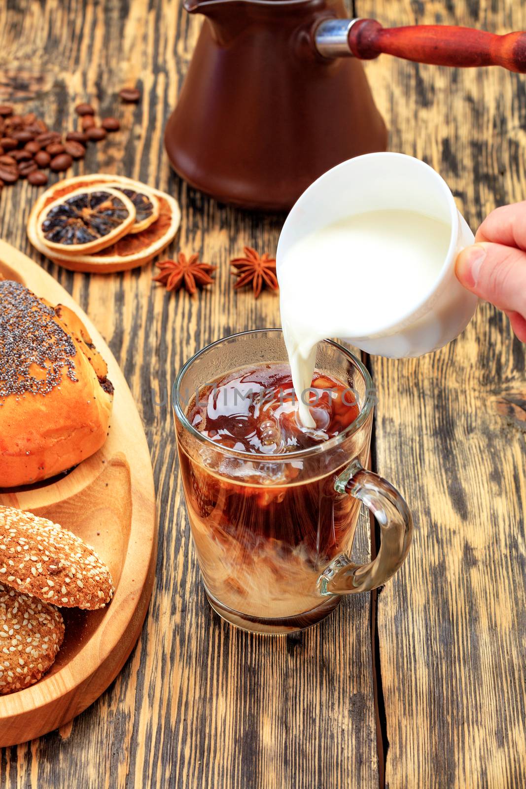 In freshly made espresso coffee in a transparent glass cup, cream is poured from a white cup against on the background of an old wooden table with coffee beans, dried orange and lemon, aniseand clay teapot in blur.