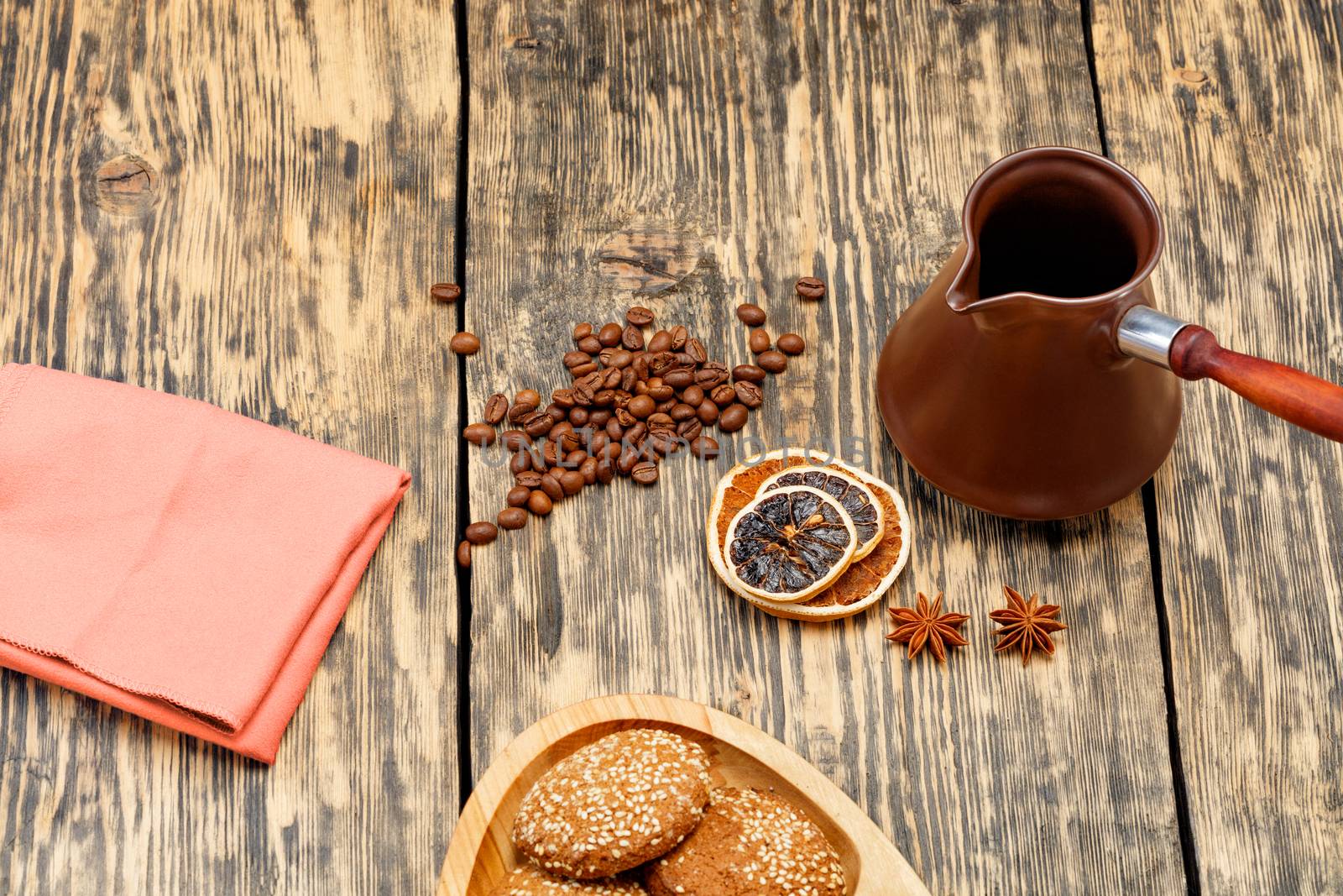 Coffee beans are scattered on an old wooden table next to dried slices of orange and lemon, anise stars, next to a coffee teapot and a light brown textile napkin in slight blur.
