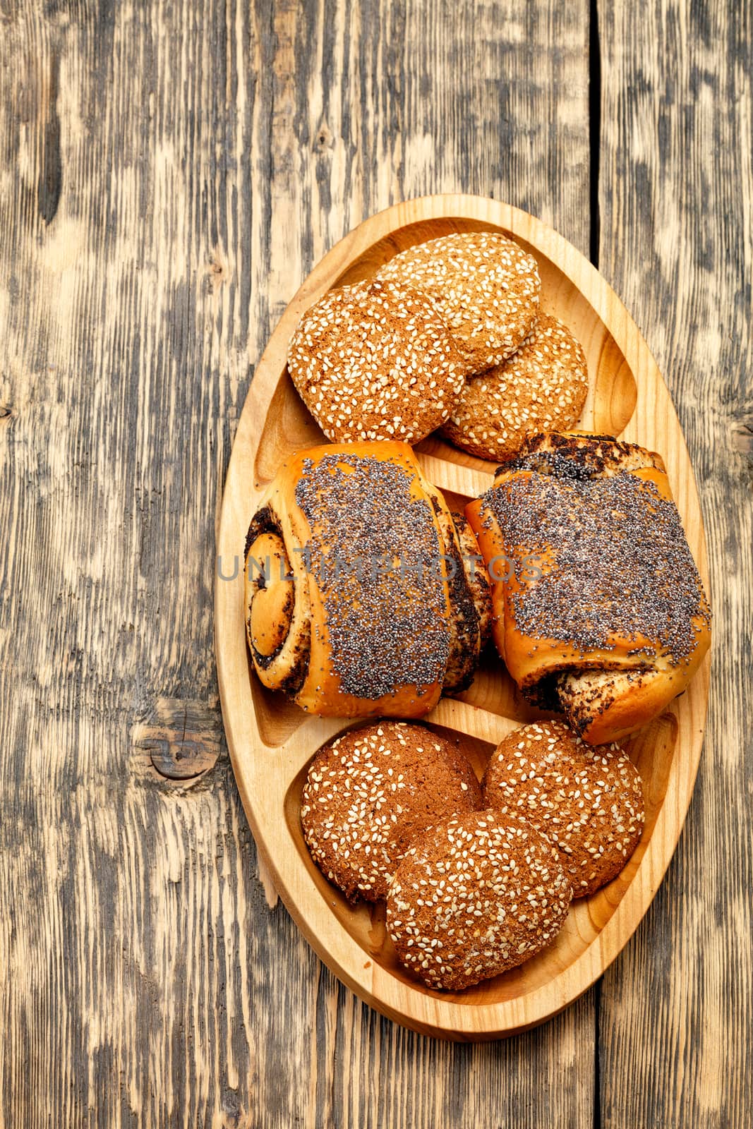 Homemade fresh buns with poppy seeds and cookies lie on a wooden oval board on a old wooden surface close-up, vertical image. by Sergii