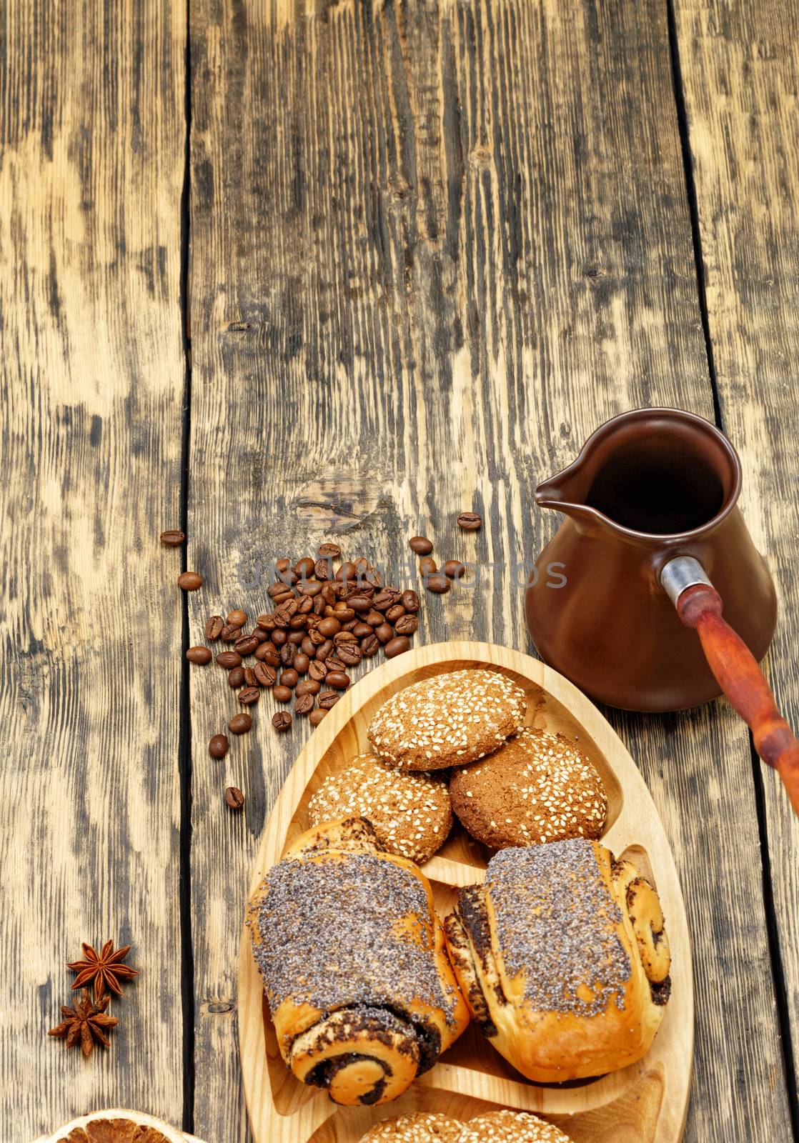 Coffee beans are scattered on an old wooden table next to dried slices of orange, anise stars, next to a coffee teapot and homemade fresh buns in slight blur.