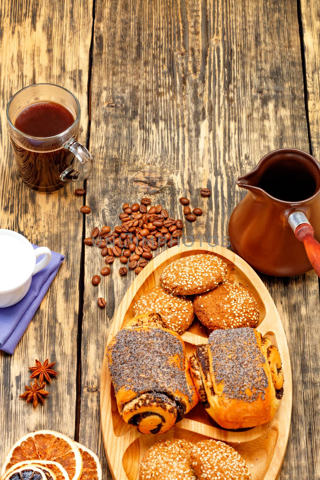 Coffee beans are scattered on an old wooden table and freshly brewed coffee in transparent glass cup to anise stars, next to a coffee teapot, cream and homemade fresh buns in slight blur.