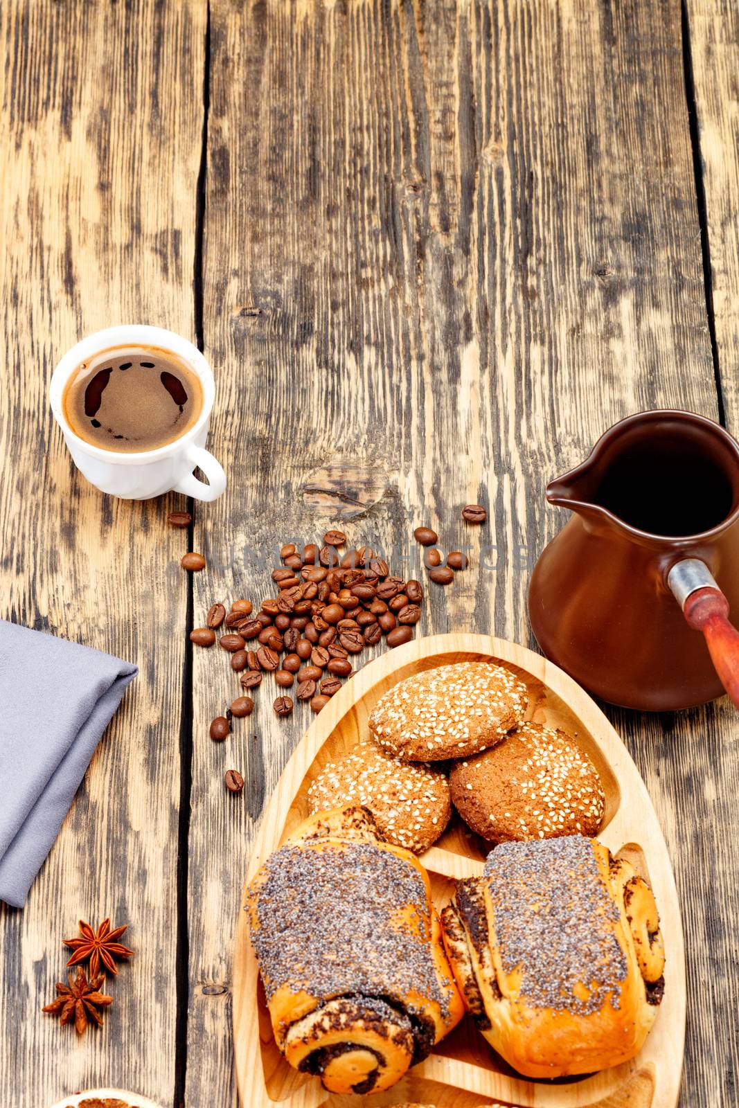 Coffee beans are scattered on an old wooden table and freshly brewed coffee in a white porcelain cup to anise stars, next to a coffee teapot and homemade fresh buns in slight blur.