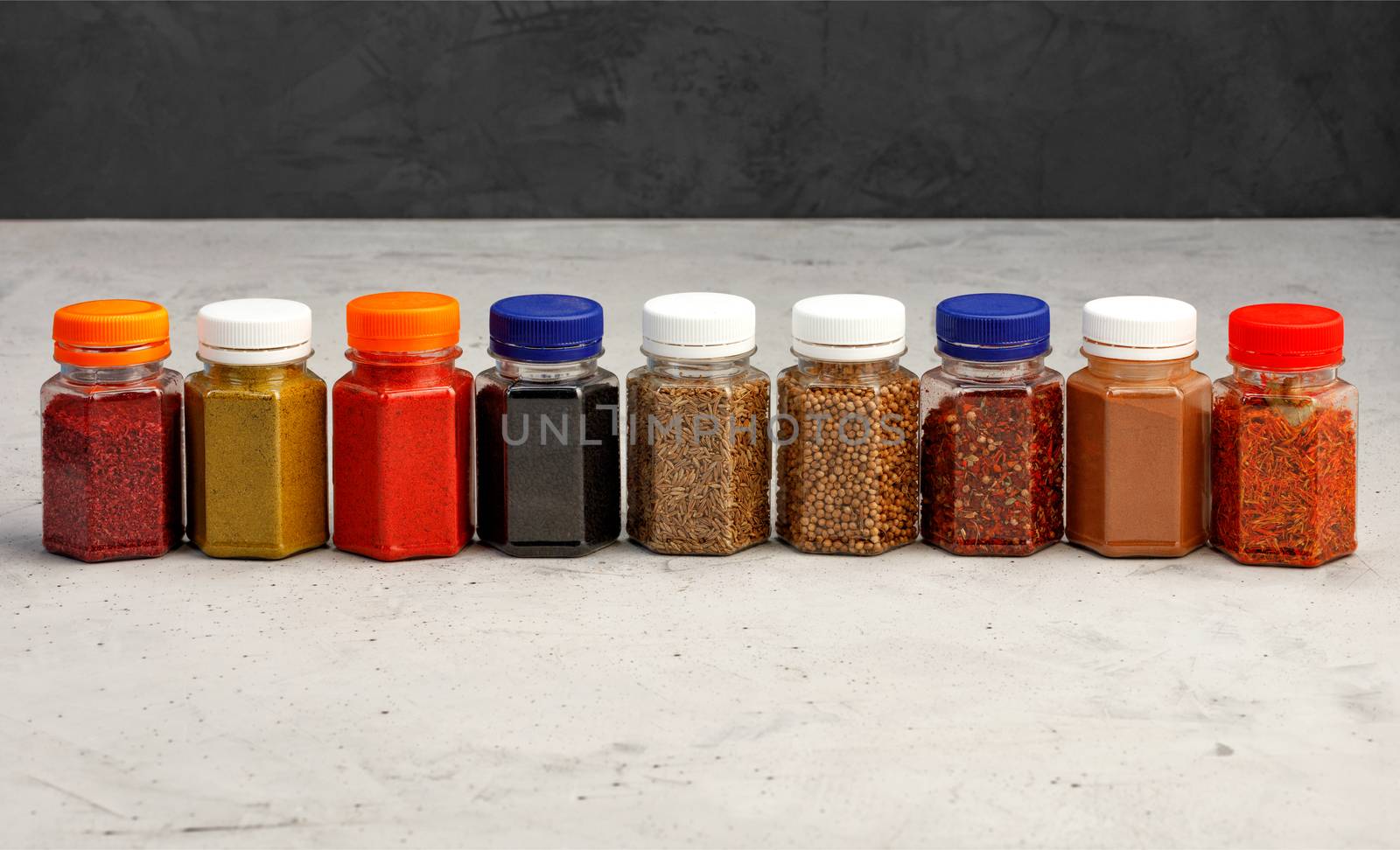 Jars glass transparent with various asian spices ground paprika, corinade, hops, zira, sumac, caraway seeds, cinnamon, saffron, allspice on a gray concrete background.