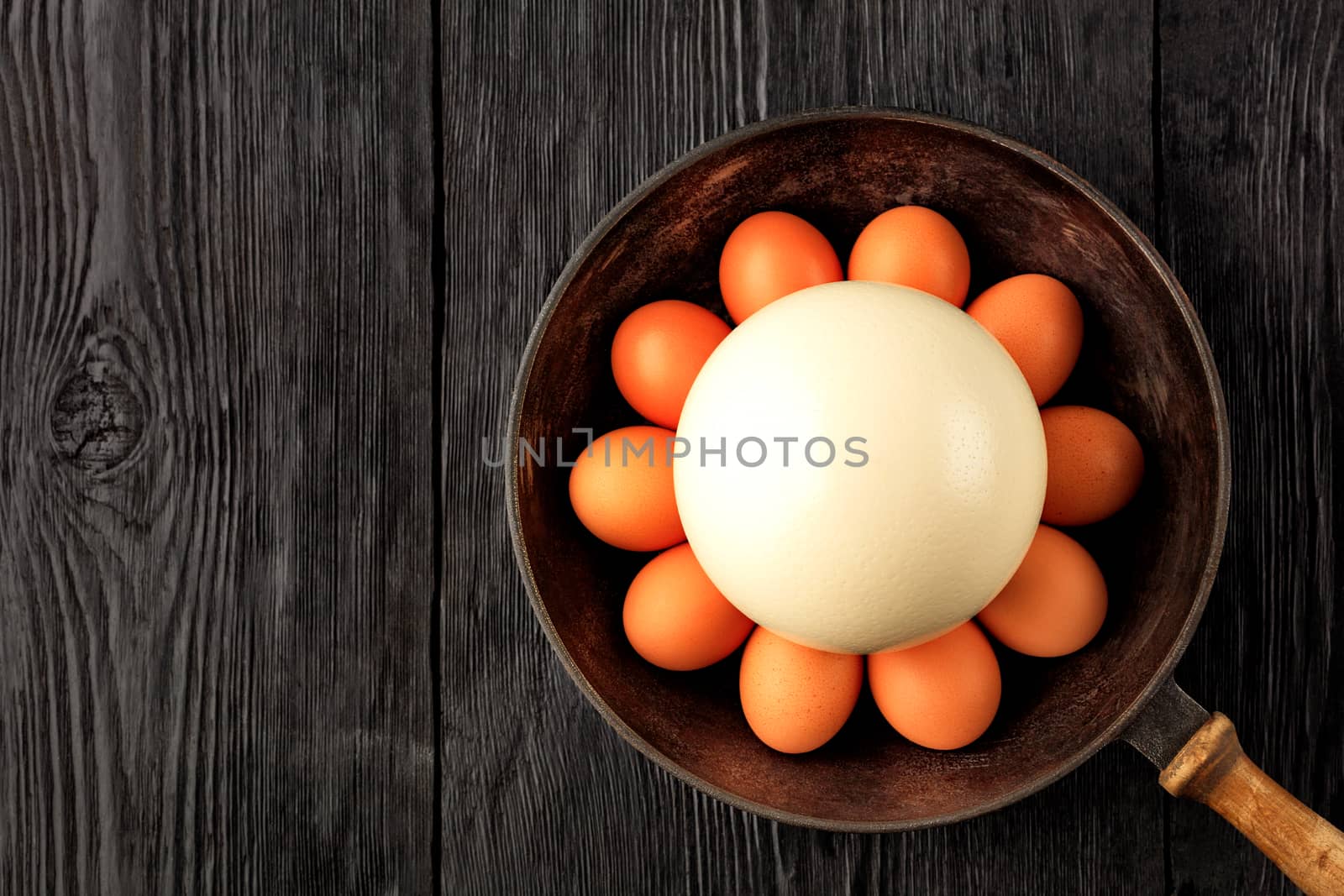 A large ostrich egg surrounded by chicken eggs in an old cast-iron pan that stands on an old black wooden surface, low key image, top view, copy space.