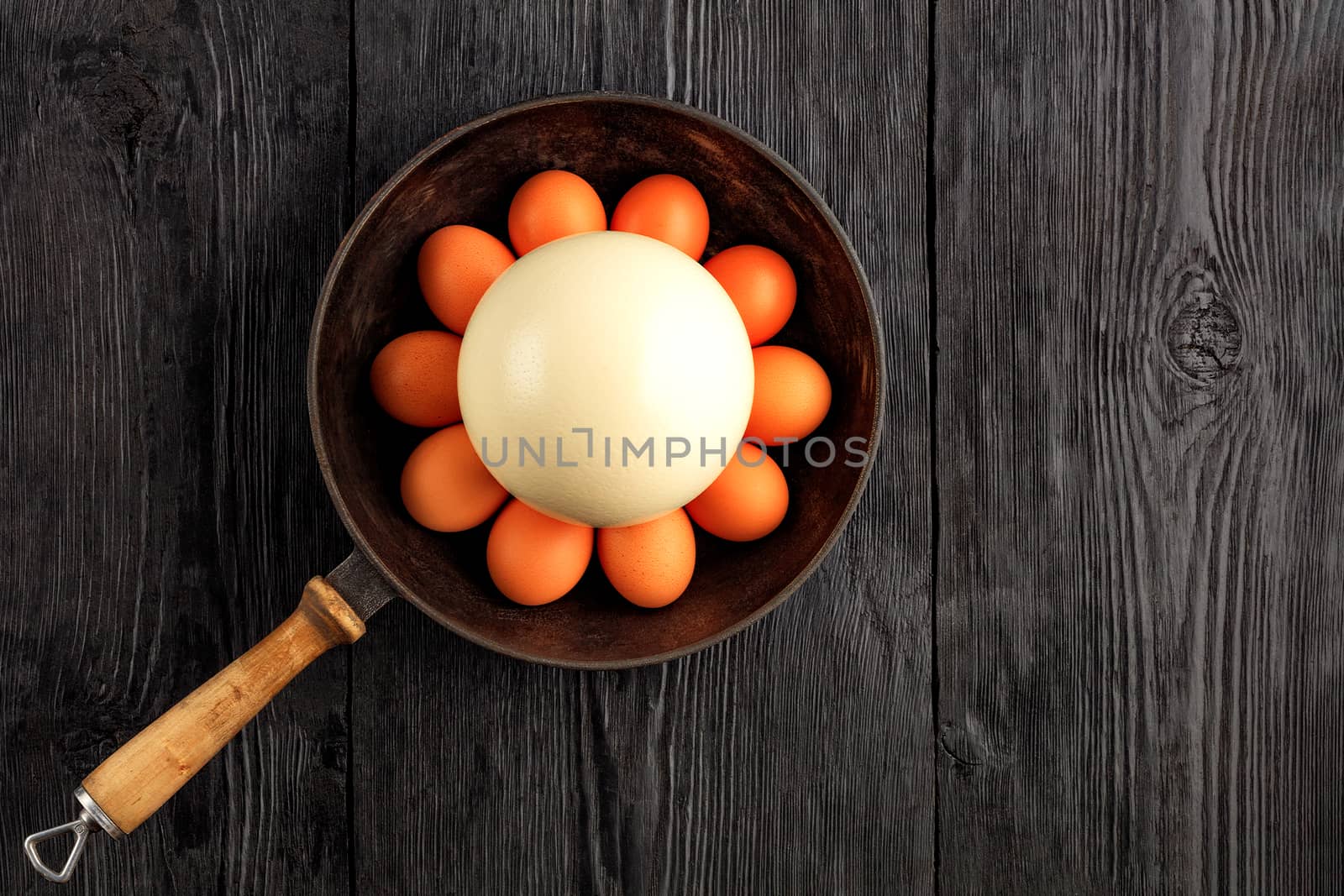 Ostrich egg surrounded by chicken eggs in an old cast-iron skillet, which is standing on an old black wooden surface, top view, copy space.
