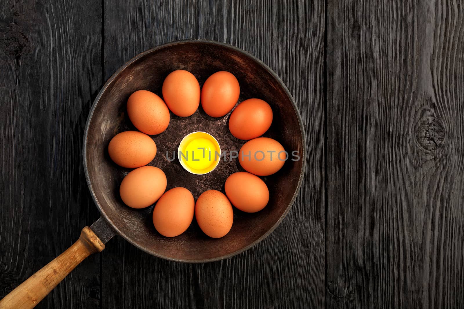 Bright chicken yolk of a broken egg surrounded by chicken eggs in an old cast-iron pan, which is standing on an old black wooden surface, top view, copy space.