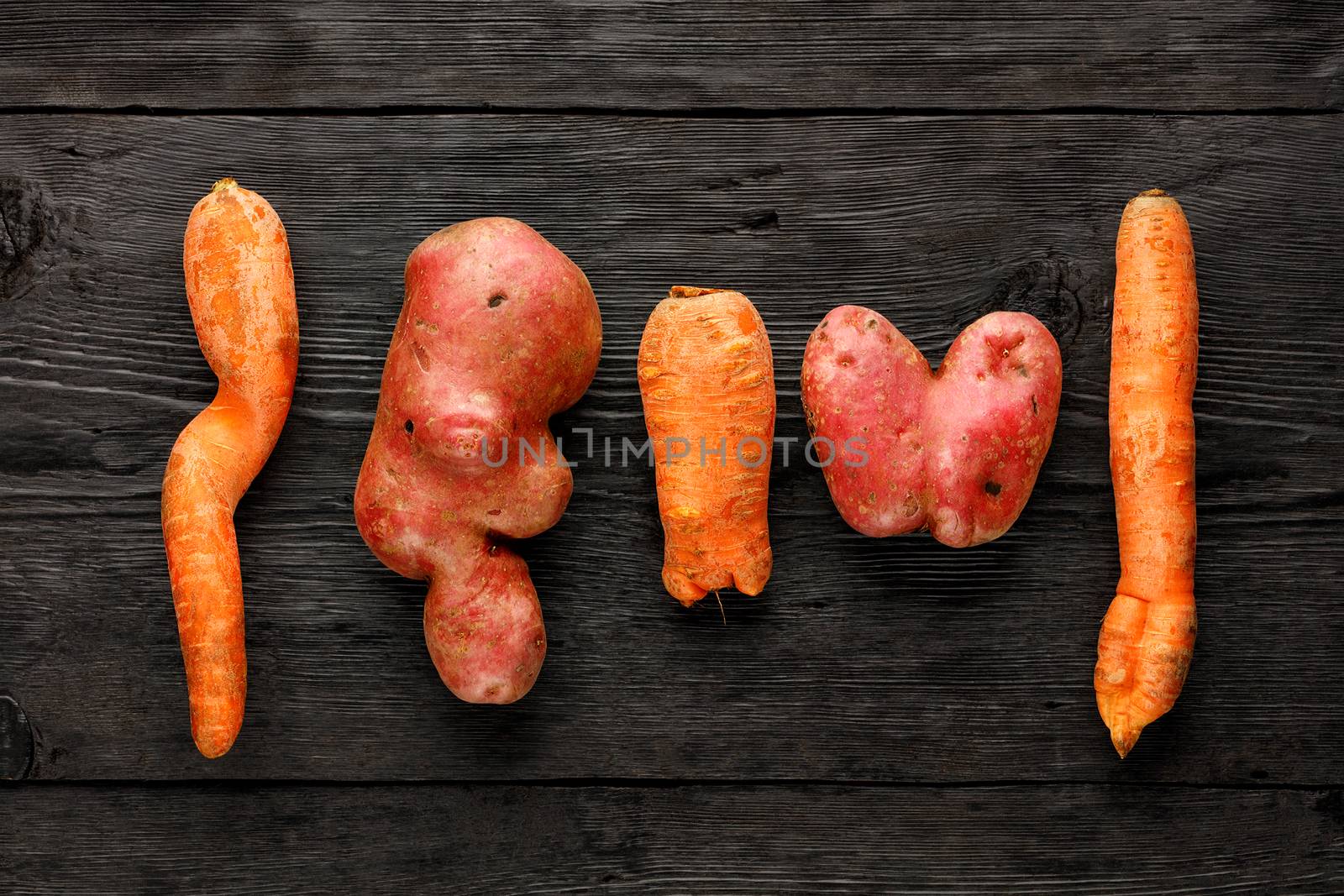 Ugly funny vegetables, heart-shaped potatoes and twisted carrot on a black wooden background. The concept of grungy vegetables or food waste. Top view, low key, copy space.