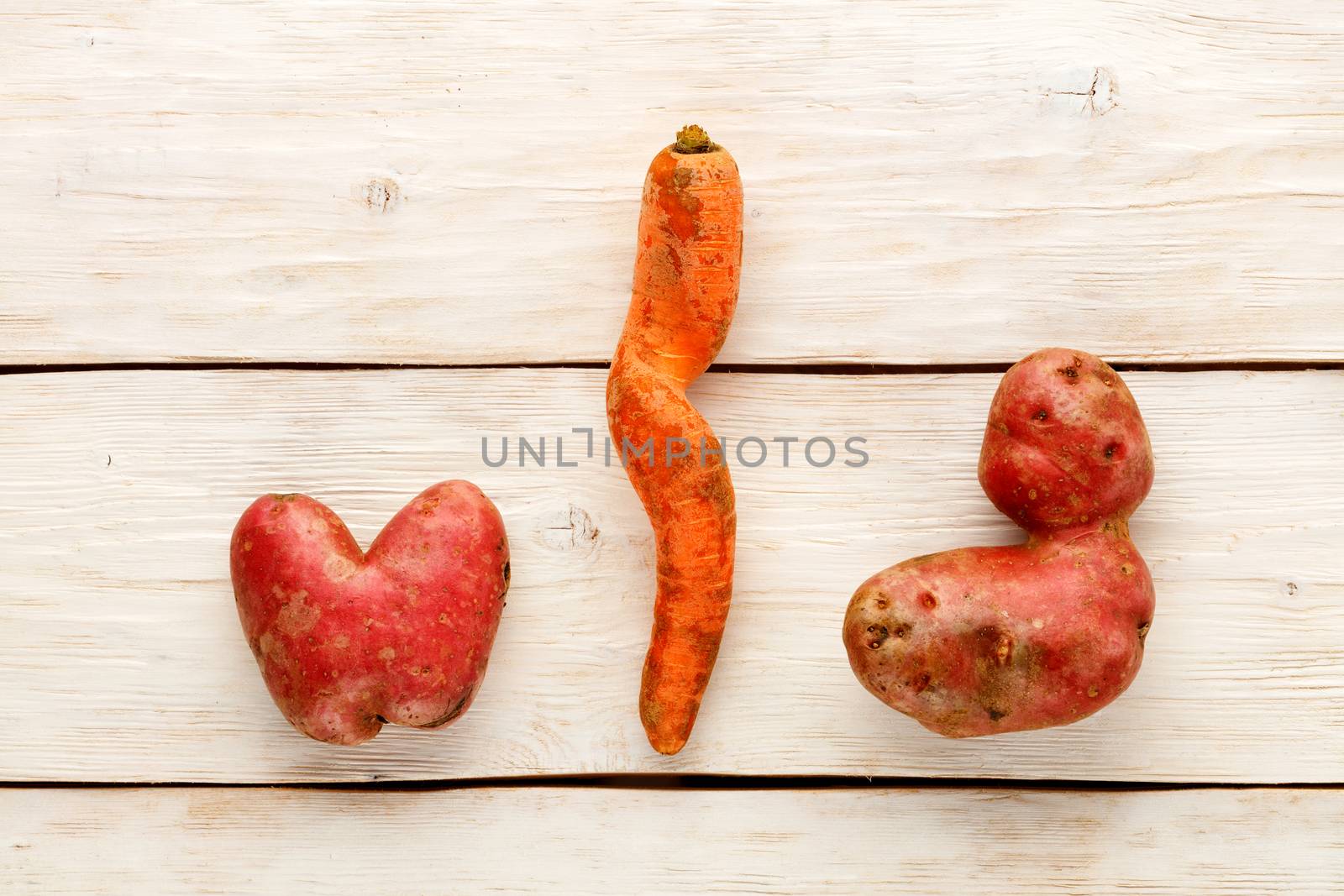Ugly funny vegetables, heart-shaped potatoes and twisted carrot on a white wooden background. The concept of grungy vegetables or food waste. Top view, copy space.