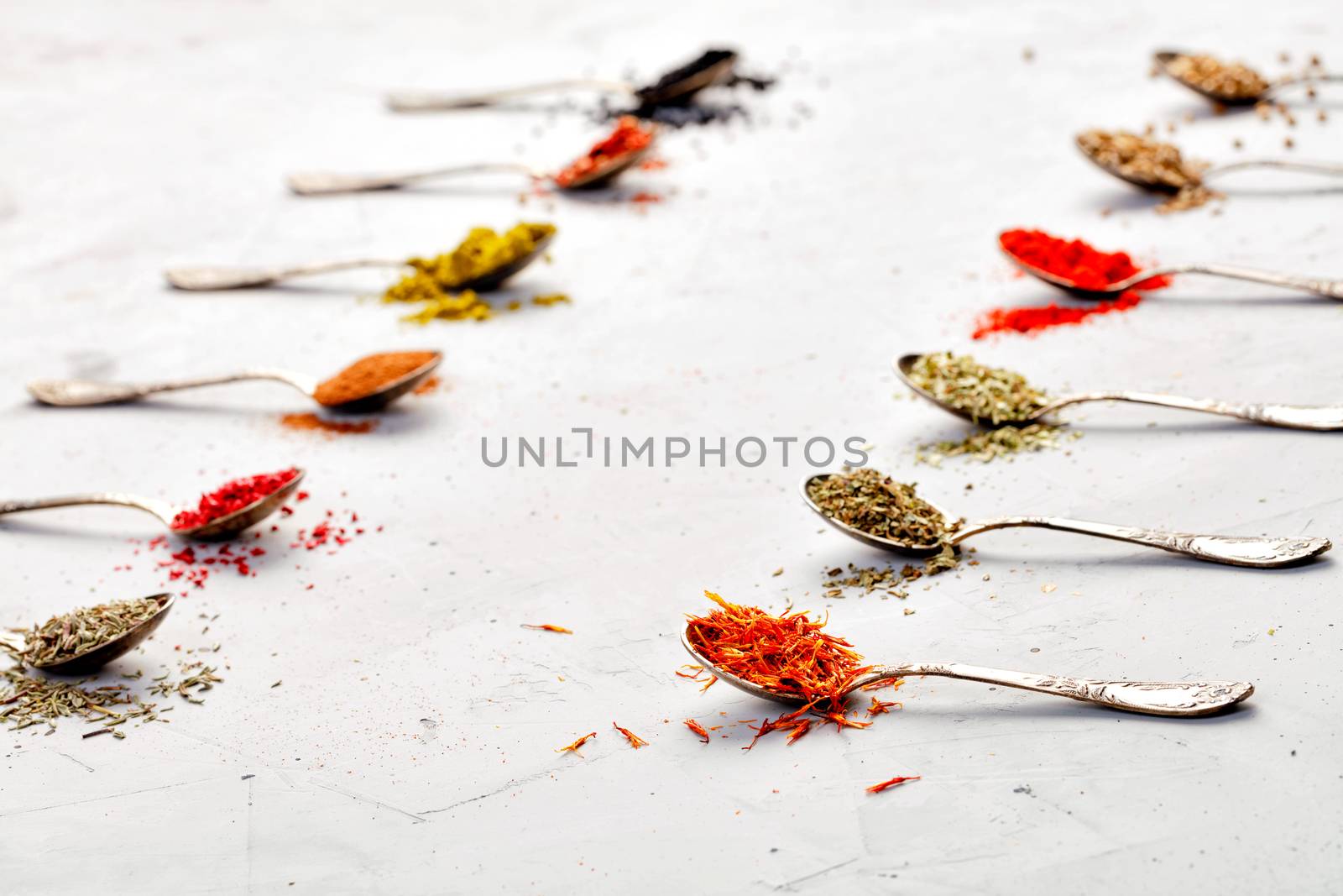 Various Asian spices saffron, zira, corinade, ground paprika, mayran, oregano are located diagonally in old silver spoons on a gray concrete background.