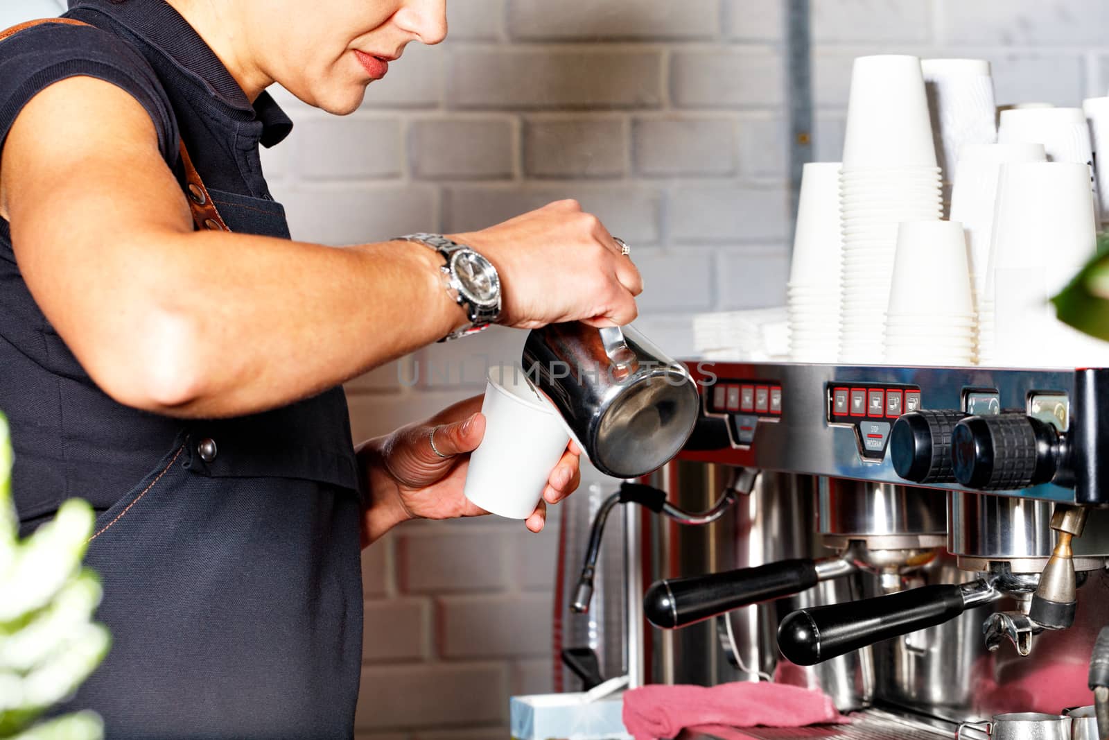 A barista in a black apron pours freshly prepared coffee from a metal mug into a paper cup against the background of a coffee machine and a stack of empty paper cups in blur.