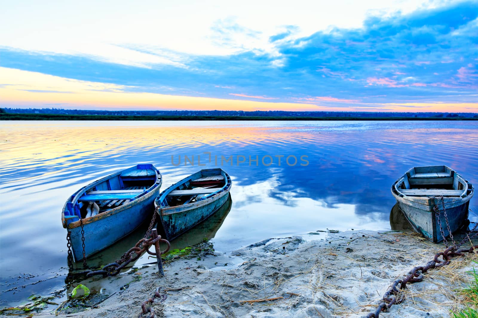Old blue-green wooden boats are chained with iron chains to the banks of a calm river. The early mornings and rays of the rising sun illuminate the horizon and summer landscape of the banks of a wide river.