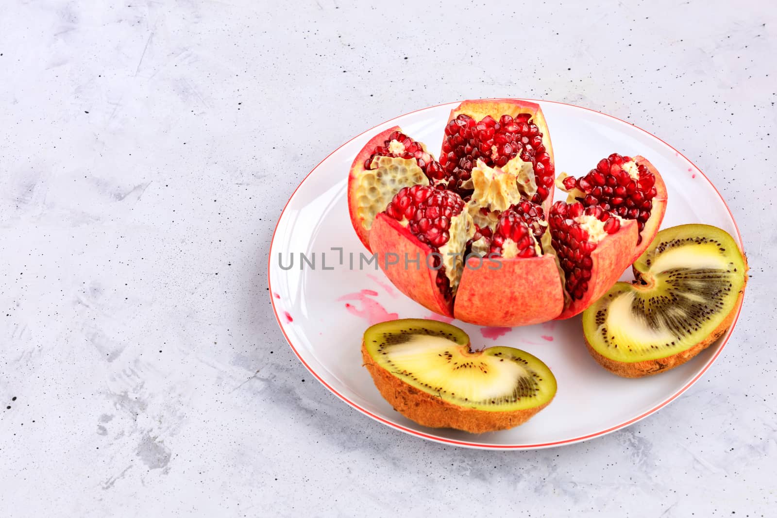 Ripe, red pomegranate fruit is divided into parts and a kiwi cut in half on a white porcelain plate, on a background of gray concrete, close-up, copy space.