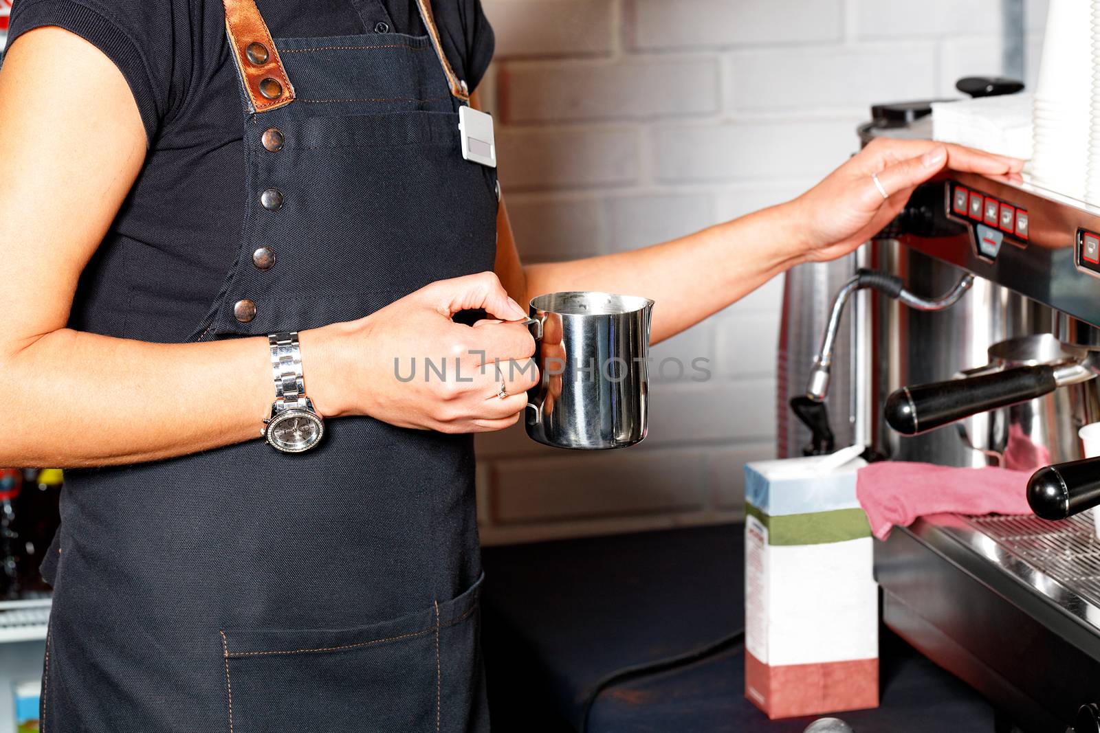 The barista in a black apron holds in his hand a metal mug for making coffee latte on the background of a coffee machine and a packet of cream in blur.