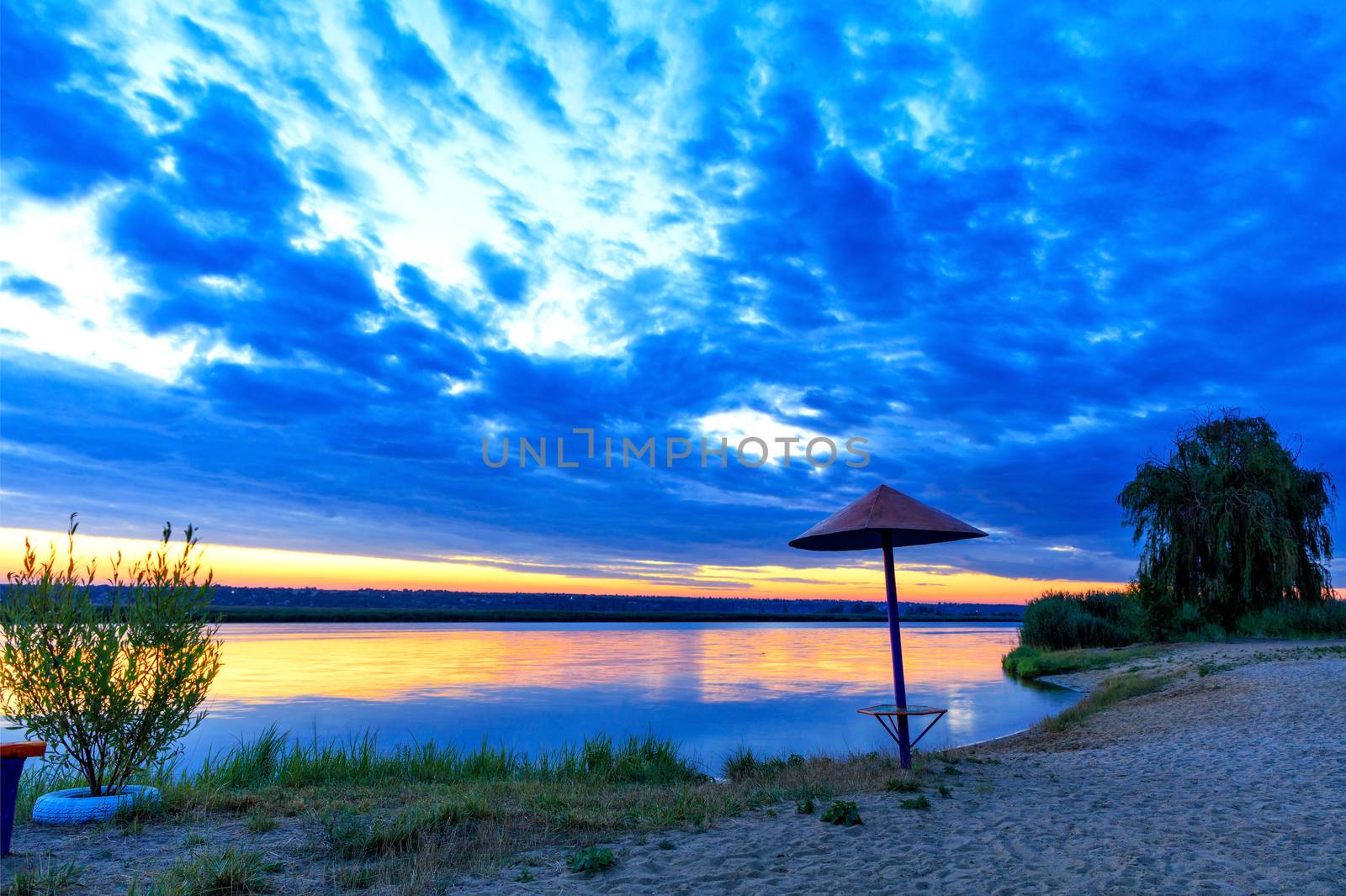 The bright golden reflection of the rays of the rising sun in the river water illuminates the coastal grass, an old wooden canopy and bushes growing on the banks of the river.