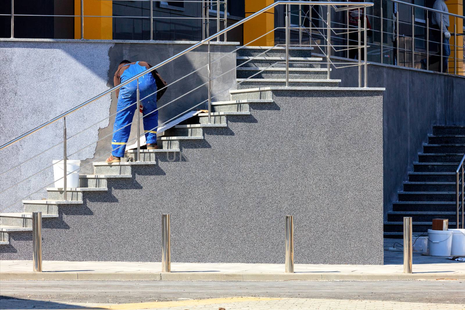 A worker in blue overalls is covering the facade of a staircase of a wall of a new house with decorative stucco on a bright sunny day, image with copy space.