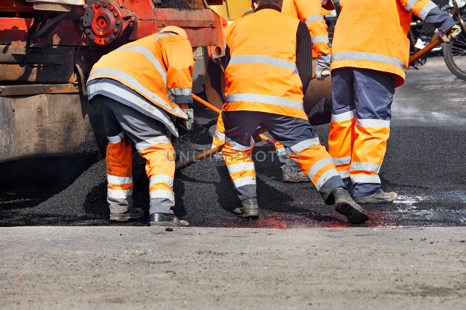A working team of road workers in an orange uniform scatters a part of the asphalt with shovels to repair a section of the road. by Sergii