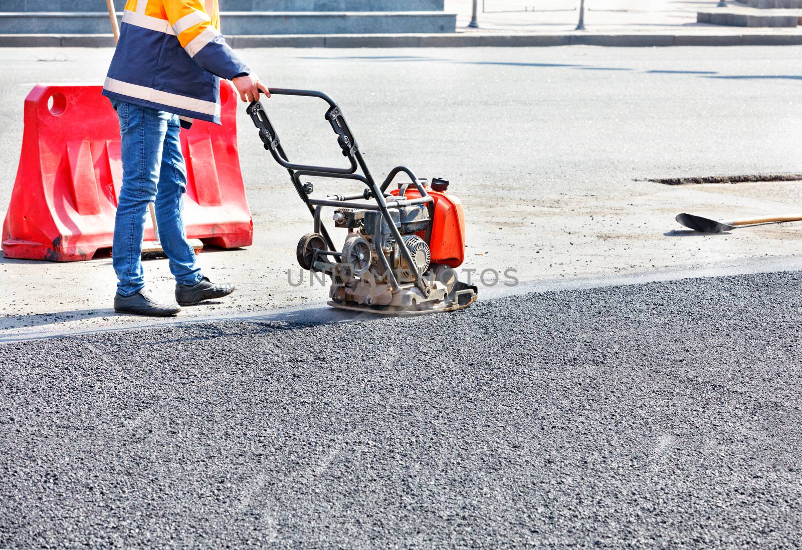 A road worker compacts asphalt in a fenced section of the road on the carriageway using a gasoline vibratory compactor.