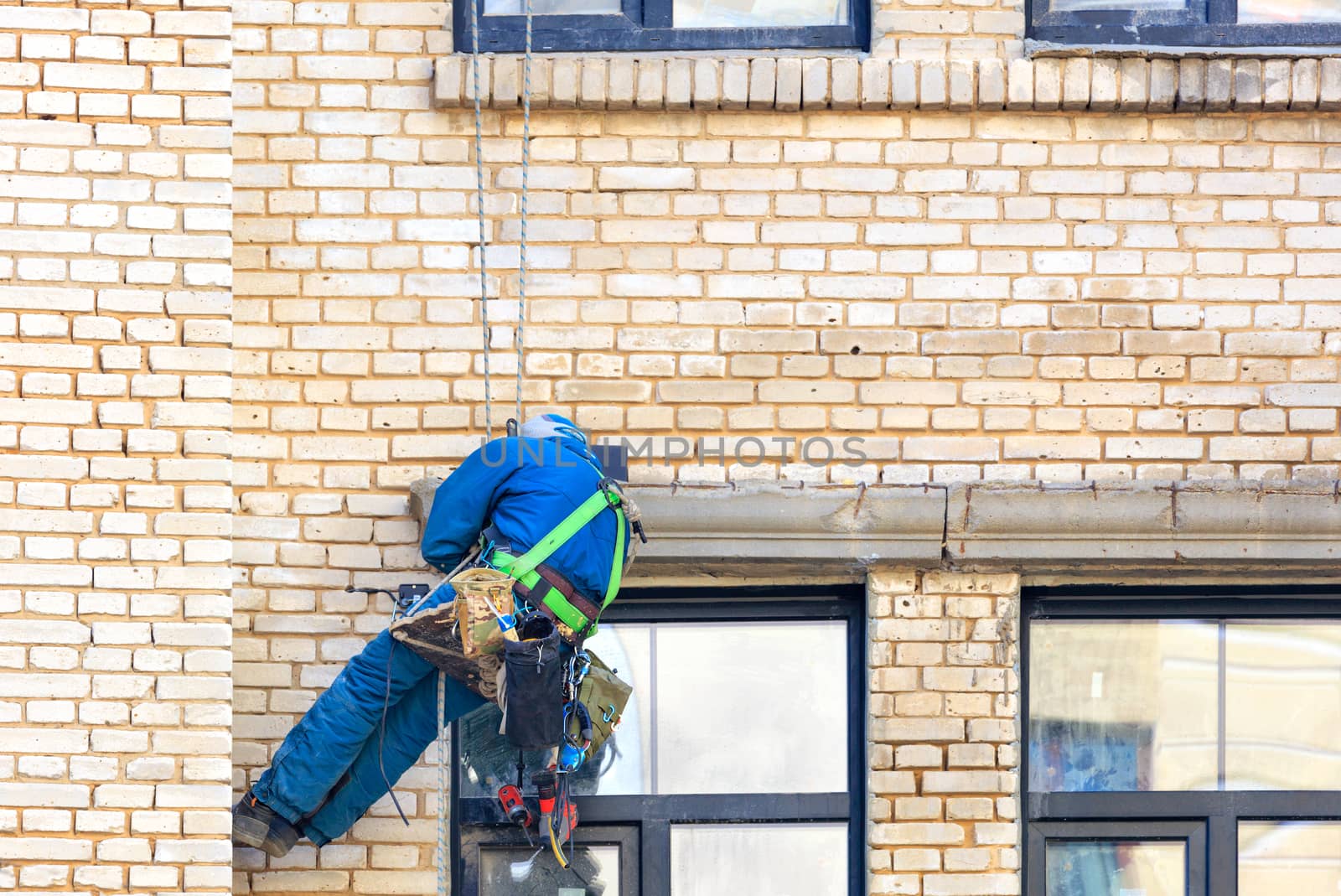 The builder of an industrial climber hangs on safety ropes on the brick wall of an old building with a set of various tools behind his back and repairs window frames, image with copy space.