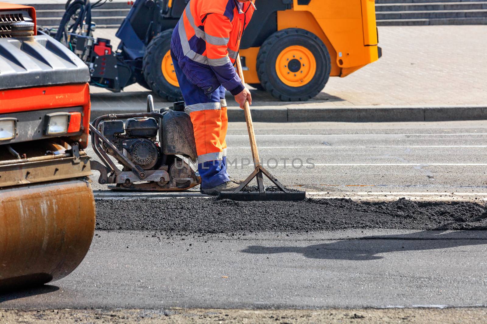 A road worker is leveling fresh asphalt on a road section against a background of road equipment. by Sergii
