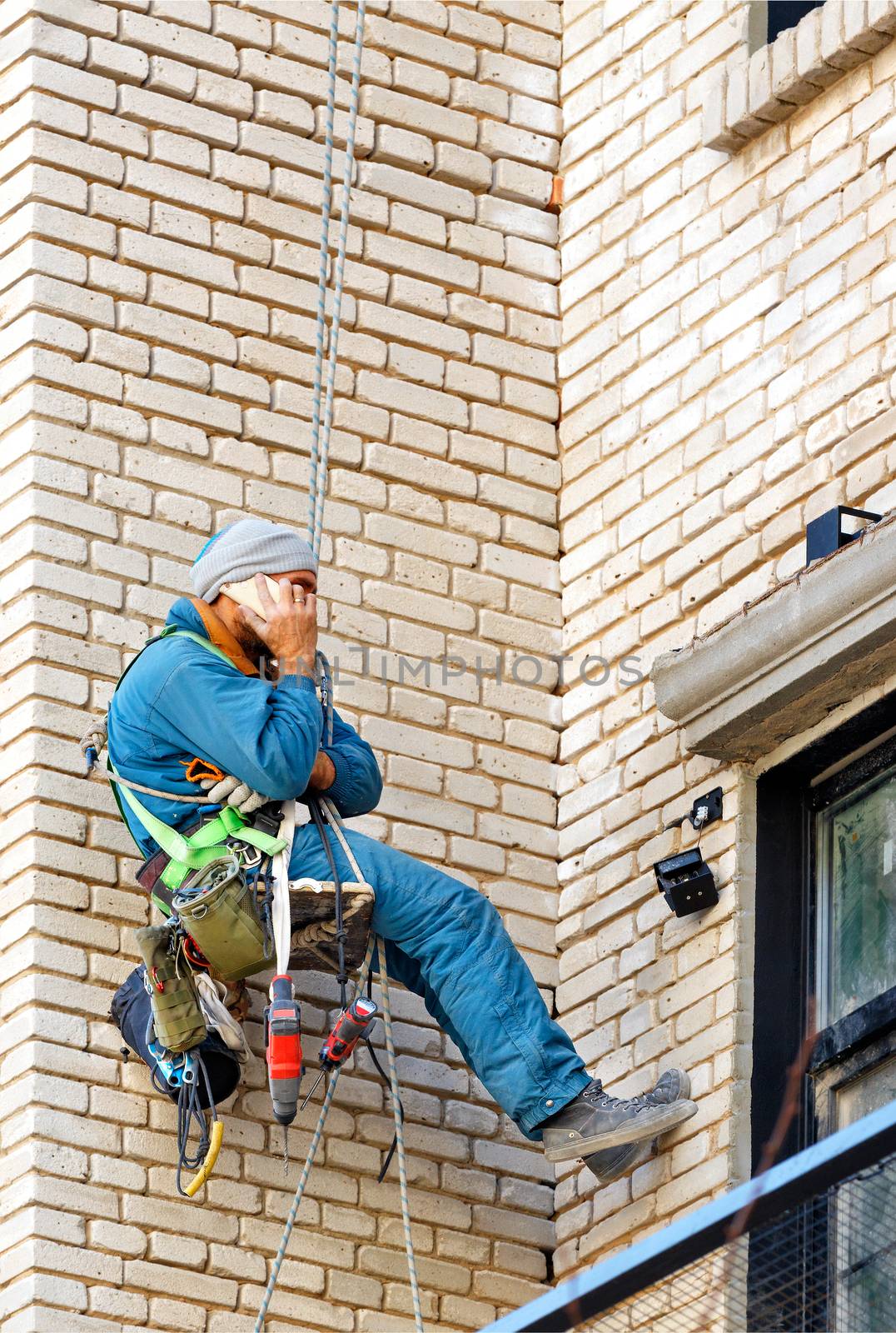 Builder climber hanging on safety ropes on the wall of an old brick building with tools tied to himself and talking on a cell phone. by Sergii