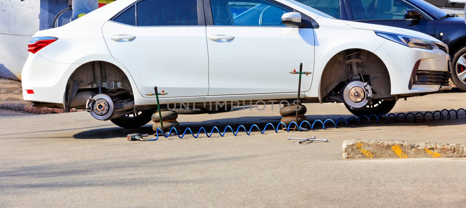 A white car without wheels stands on air jacks at a service station to replace tires.