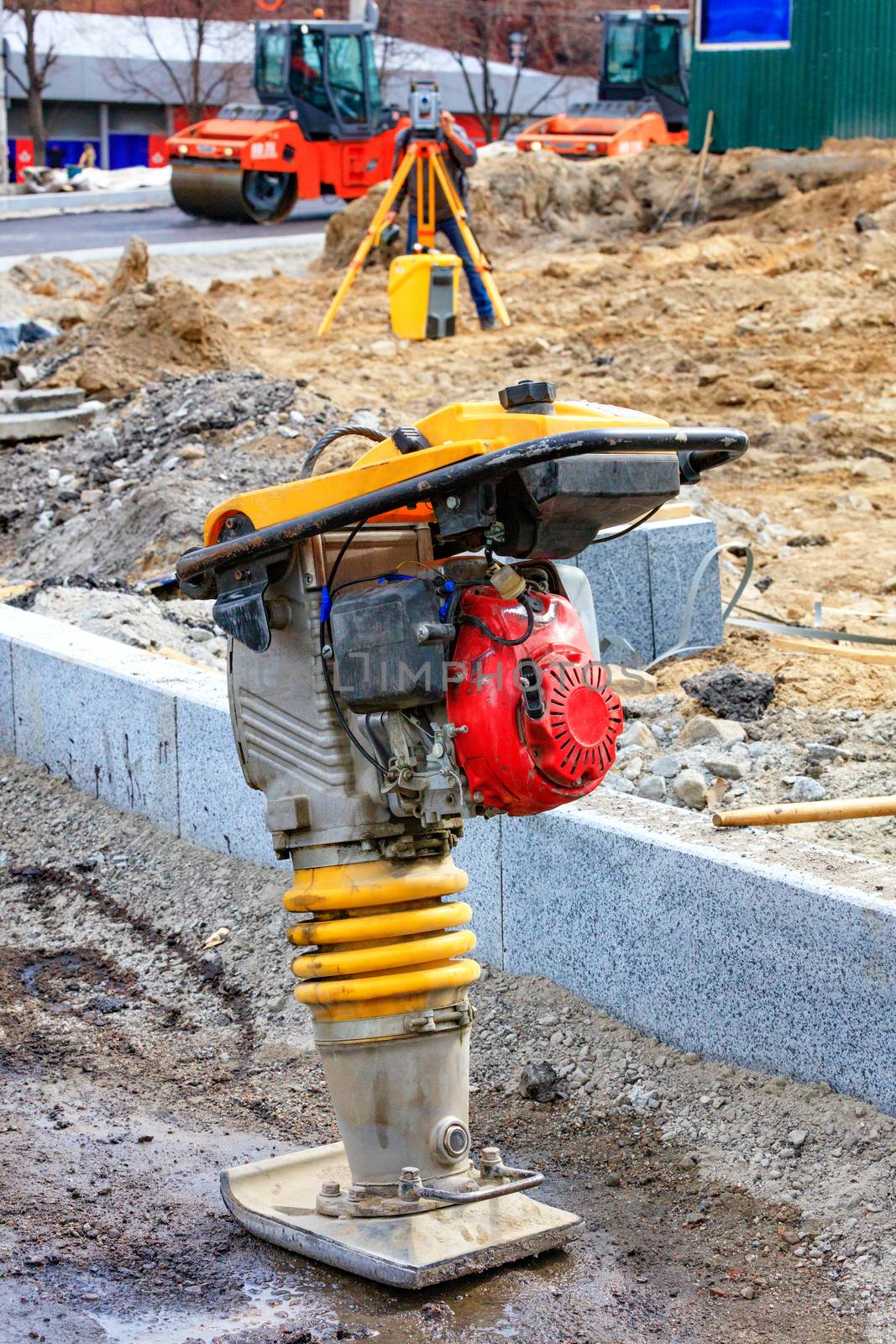 A gasoline vibratory rammer stands on a mixture of rubble and sand against the background of a road construction site in blure.