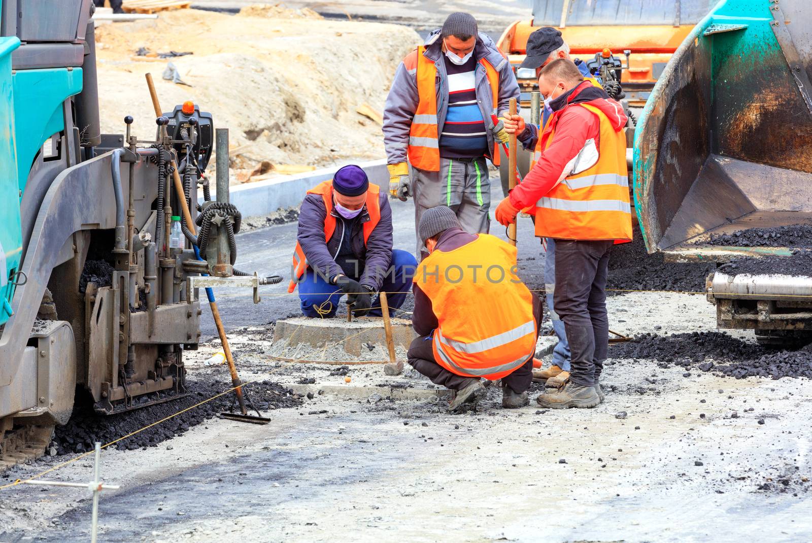 At a new work site, among large pavers, a workers team in protective masks discusses and sets the necessary level for asphalting the road.