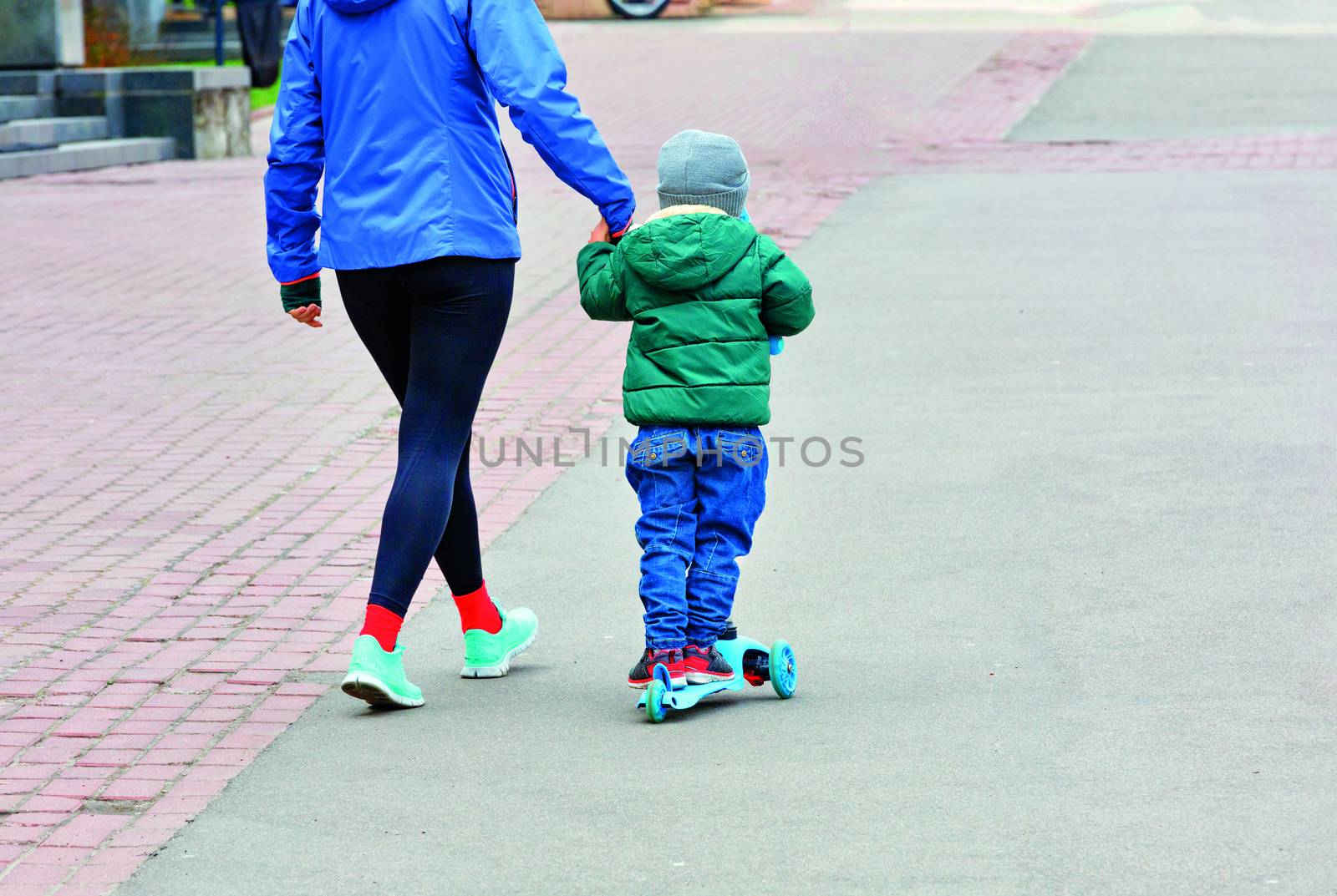 The kid in blue jeans and a green jacket on a children's scooter walks with his mother on the paved sidewalk. by Sergii