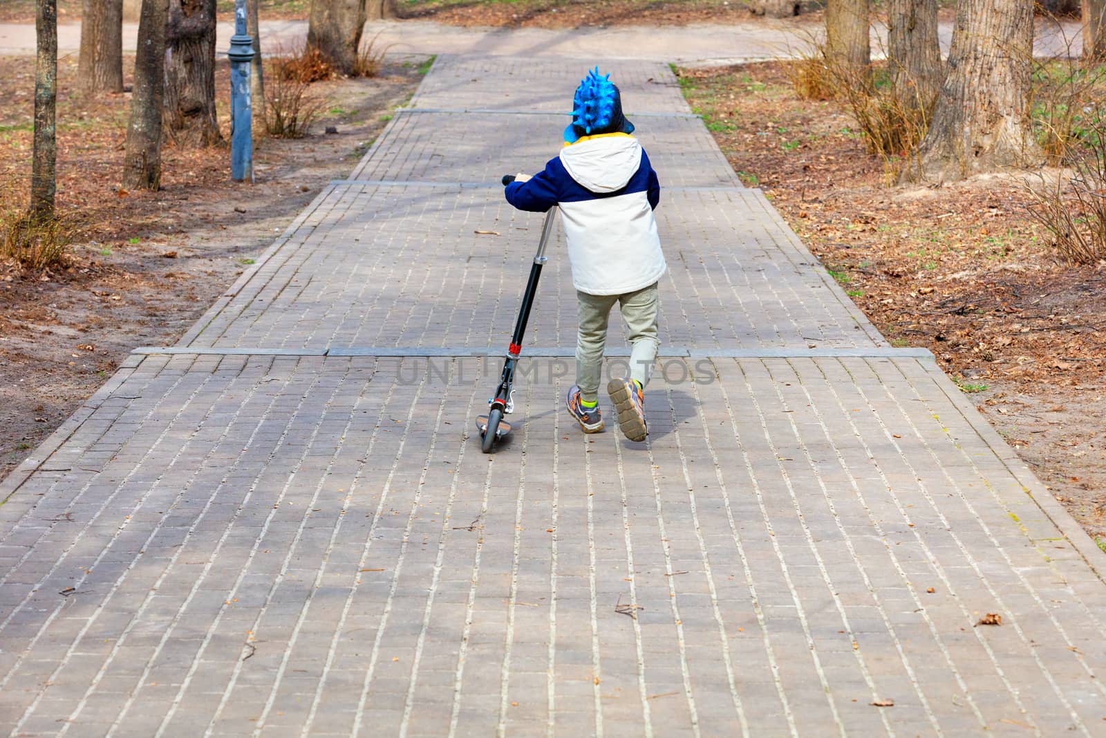 A little boy in a beige jacket and with a scooter walks in a city spring park walking along a paved path.
