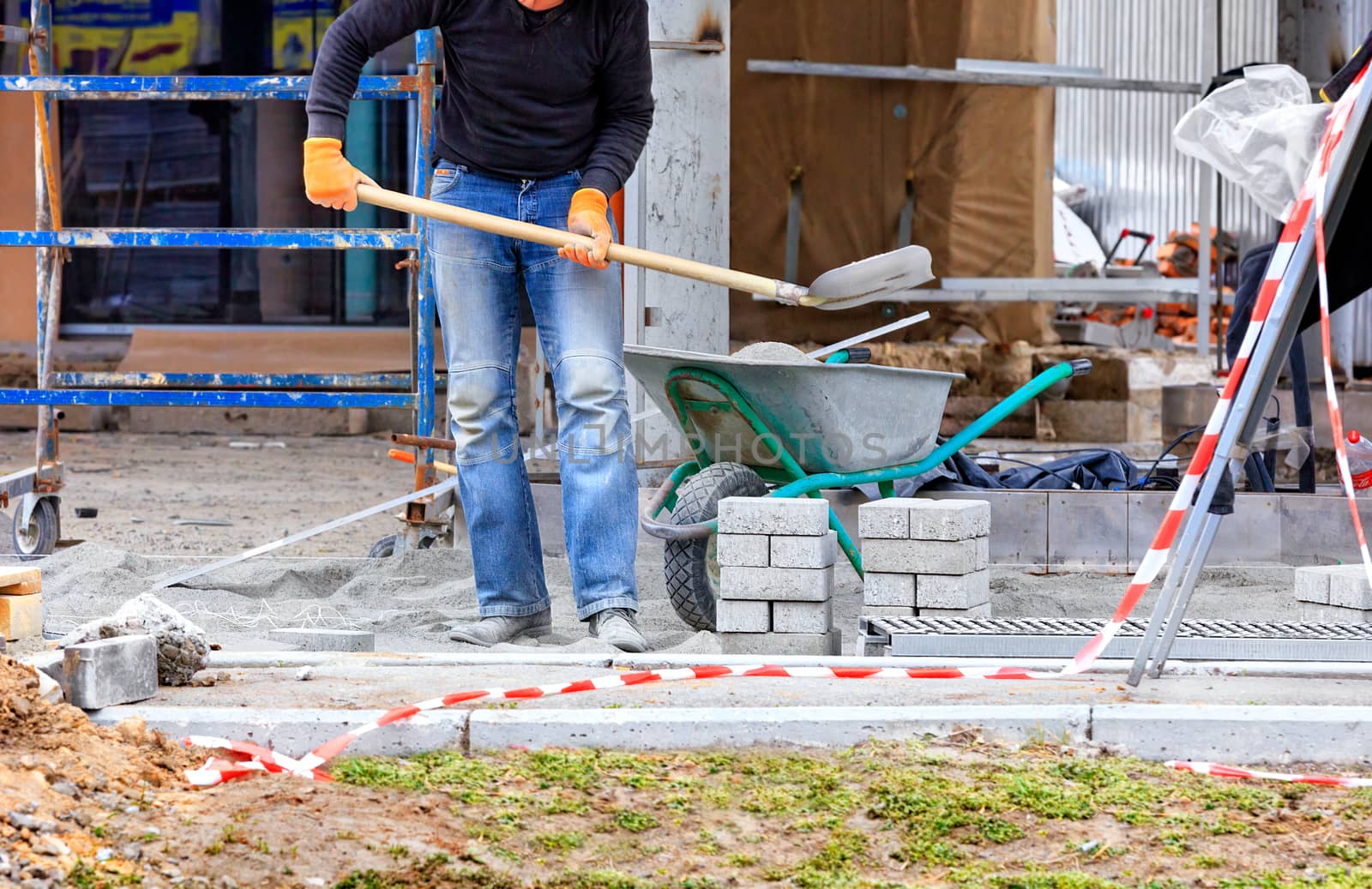 A worker levels the foundation with sand and cement using a shovel from a construction trolley for laying paving slabs against the background of a prepared site for work.