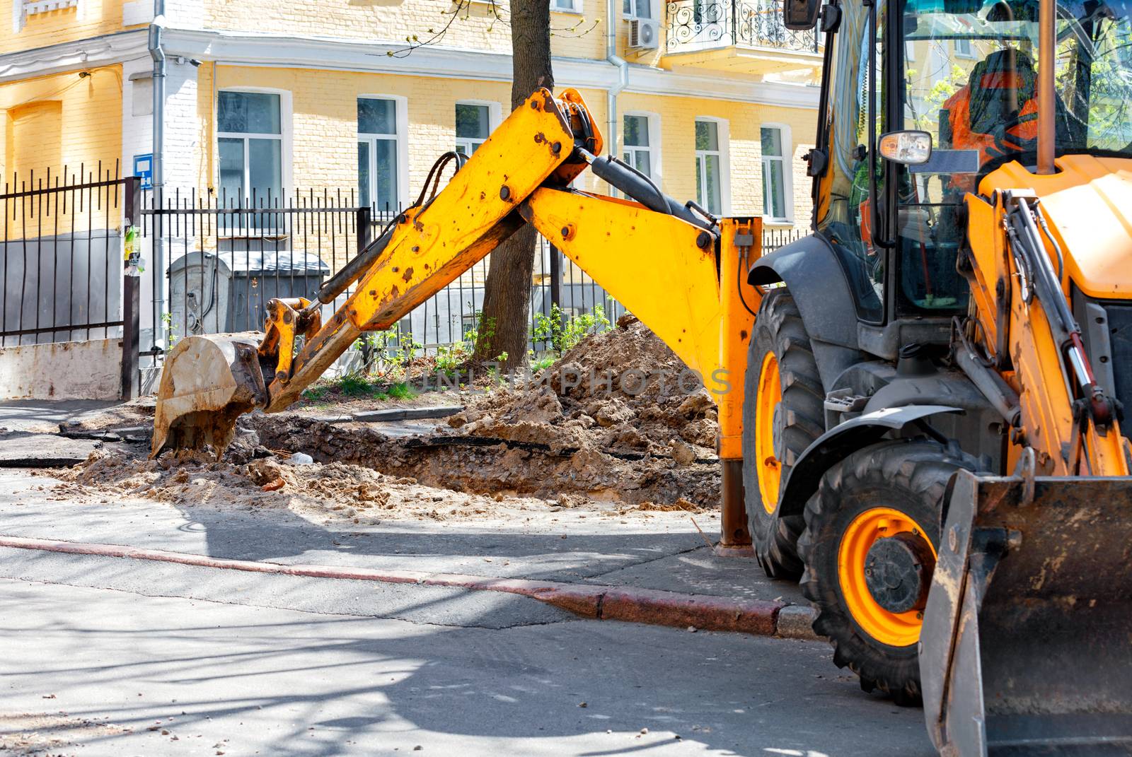 The bucket and arrow of a heavy road excavator dig a trench on the sidewalk to repair and lay urban communications.