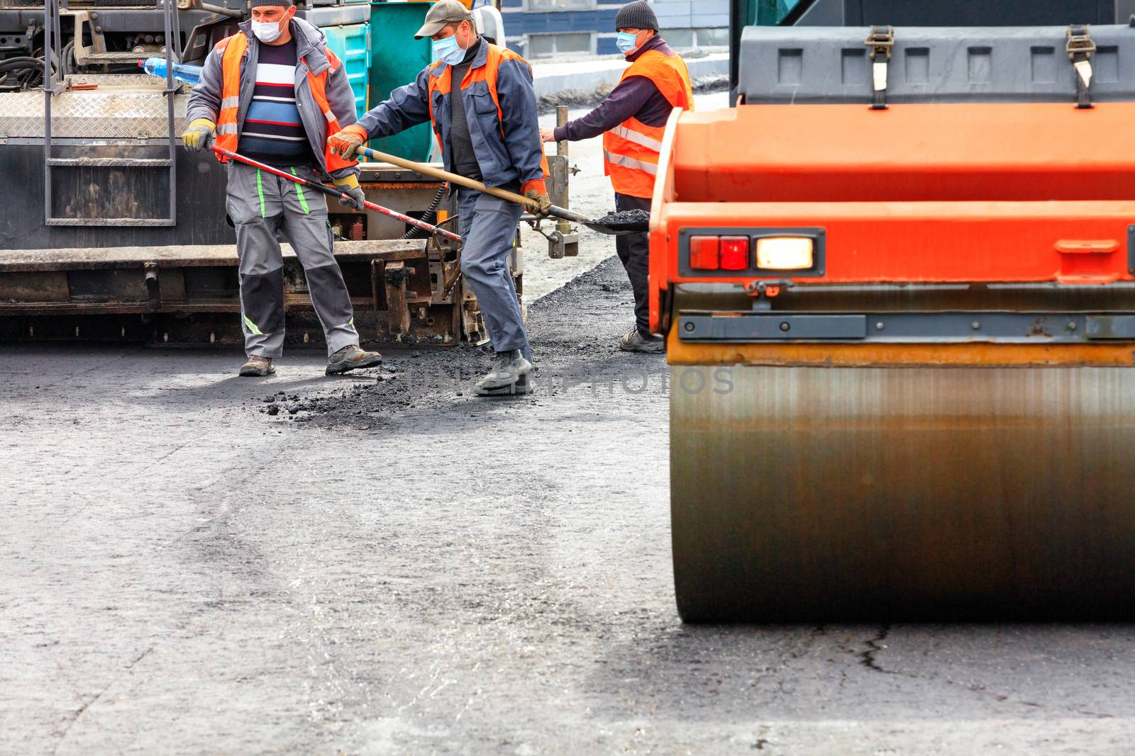 A working team of road workers in protective medical masks and overalls, using powerful road equipment and shovels, lay a new asphalt coating on the work site, image with copy space.