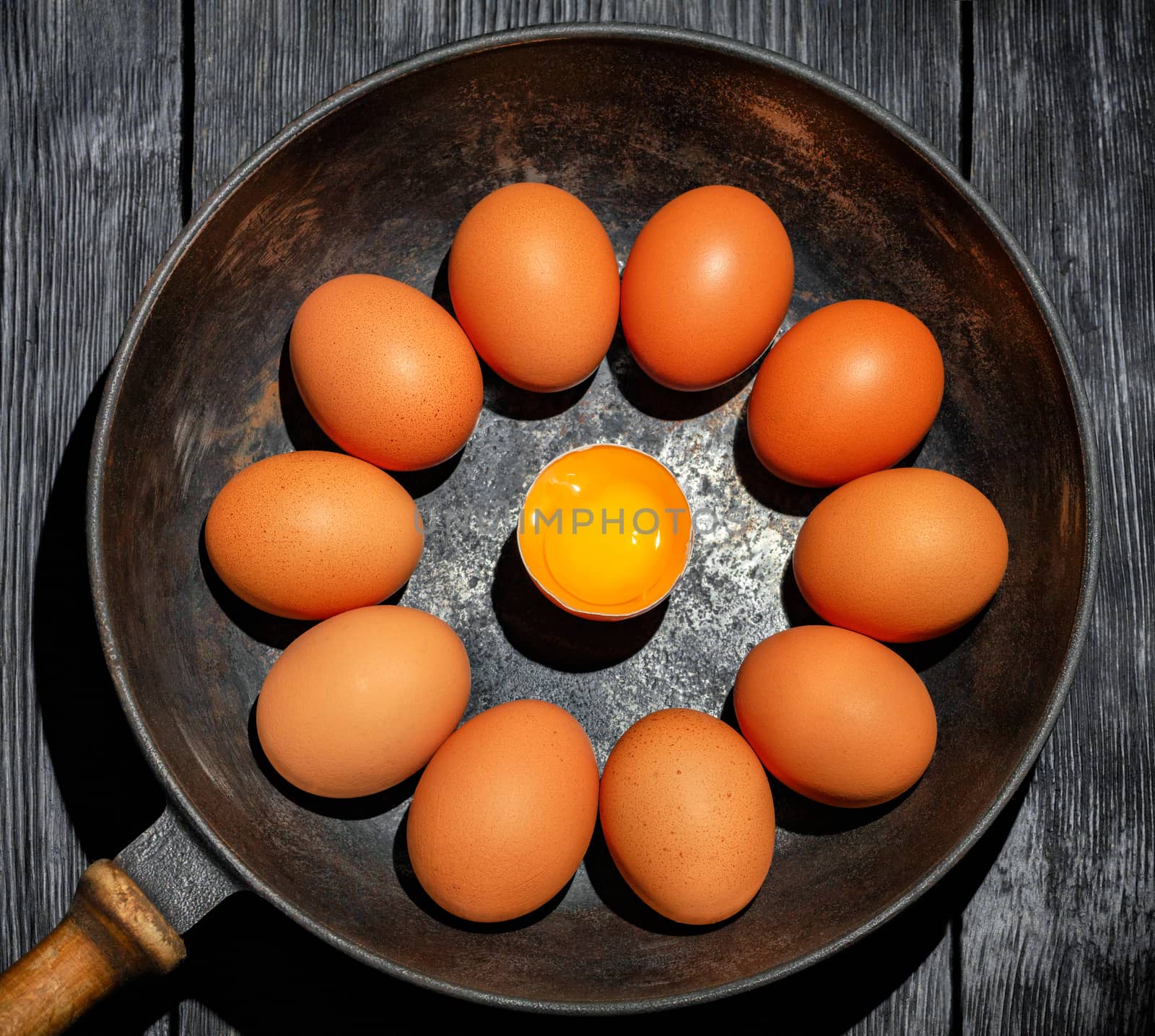 Bright chicken yolk of a broken egg surrounded by chicken eggs in an old cast-iron pan, which stands on a black wooden table, rustic style, low key, top view.