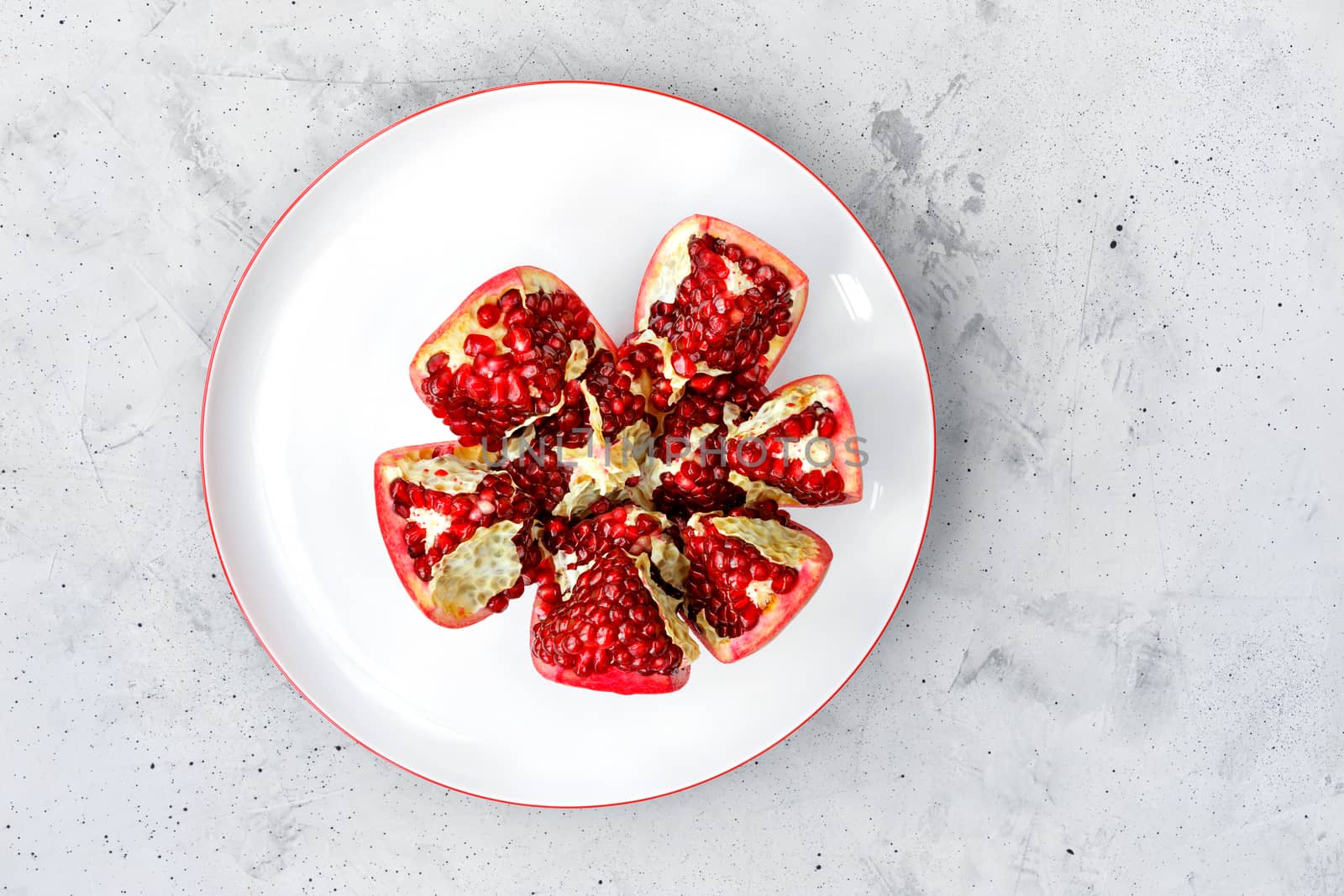 Red pomegranate fruit is divided into pieces on a white porcelain plate on a gray concrete background, copy space.