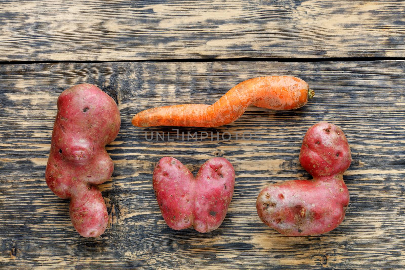 Ugly funny vegetables, heart-shaped potatoes and twisted carrot on a striped brown wooden background. The concept of grungy vegetables or food waste. Top view, low key, copy space.