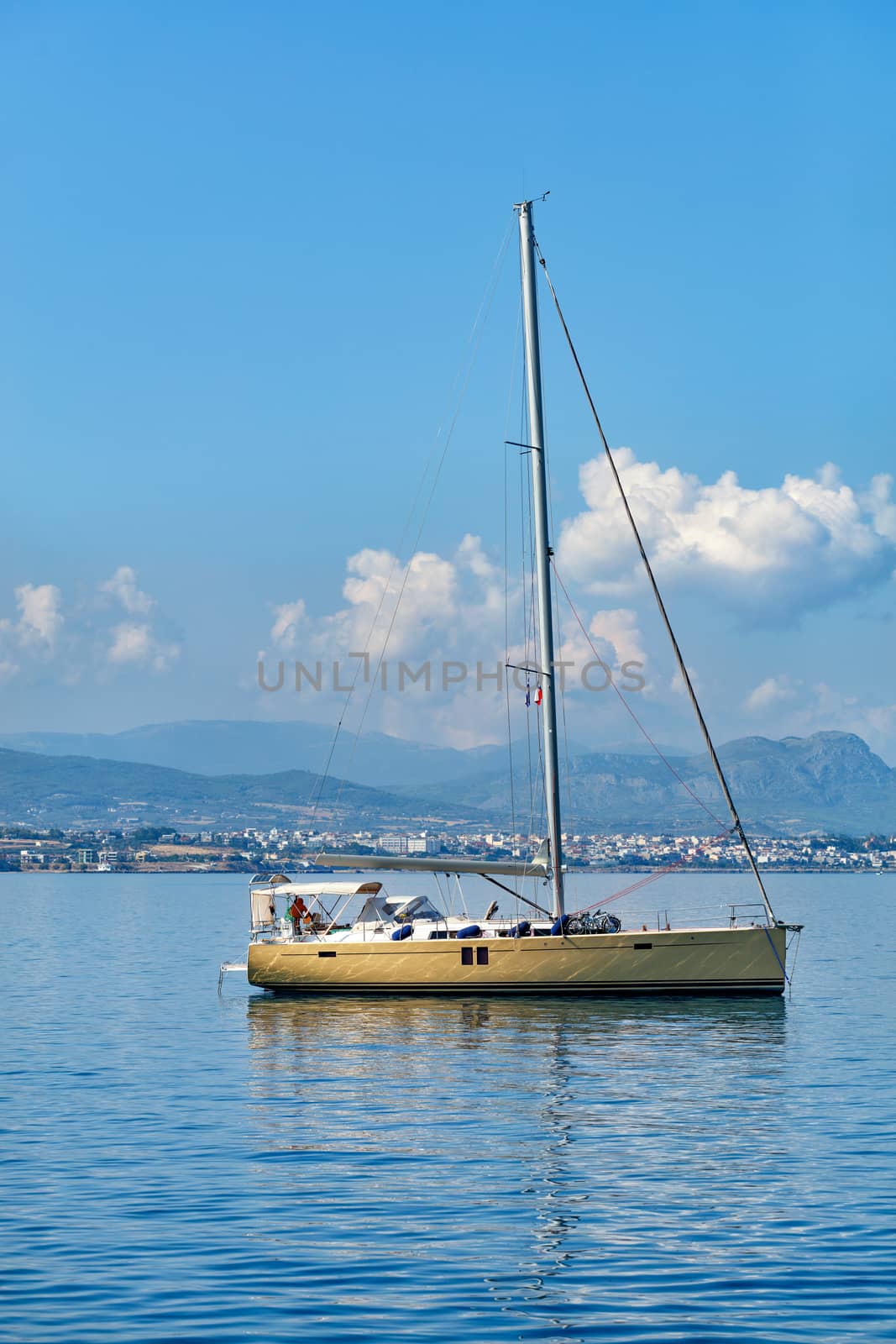 A sailing yacht is anchored against the backdrop of a mountain range and blue in the morning haze of the Gulf of Corinth and with small clouds in the blue sky.