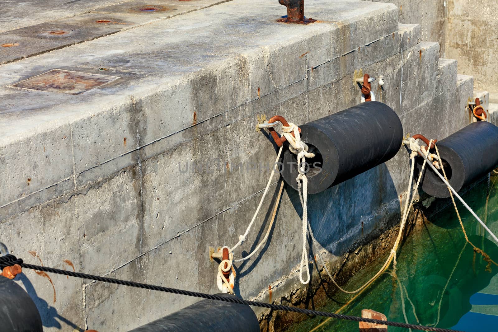 The mooring of bollards and the wing on the concrete pier of the sea pier with ropes and rusty metal brackets near turquoise and blue water.