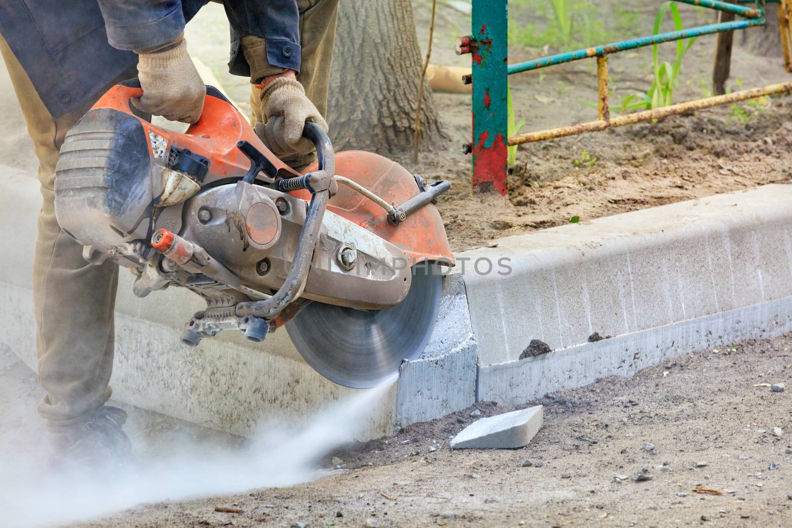 A worker using a concrete cutter cuts concrete curbs in a cloud of concrete dust in an open space.