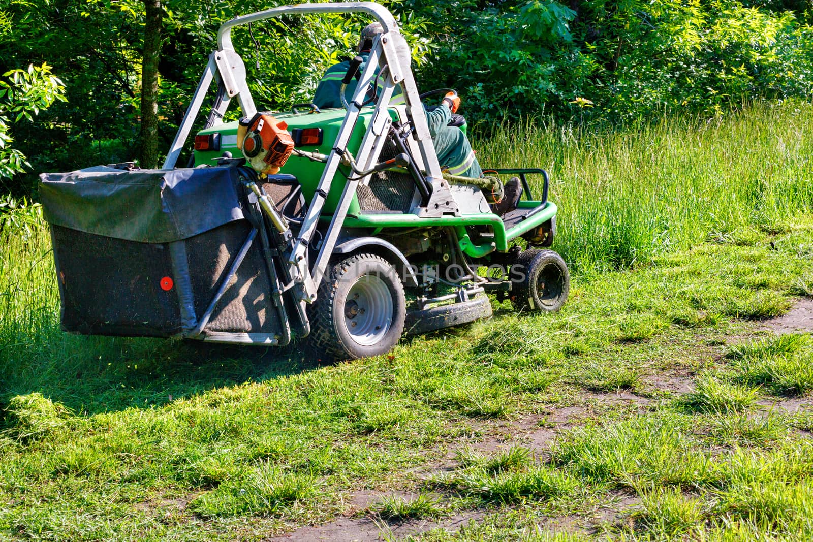 A professional tractor mower driven by a public utility worker climbs up the slope and mows tall grass. by Sergii