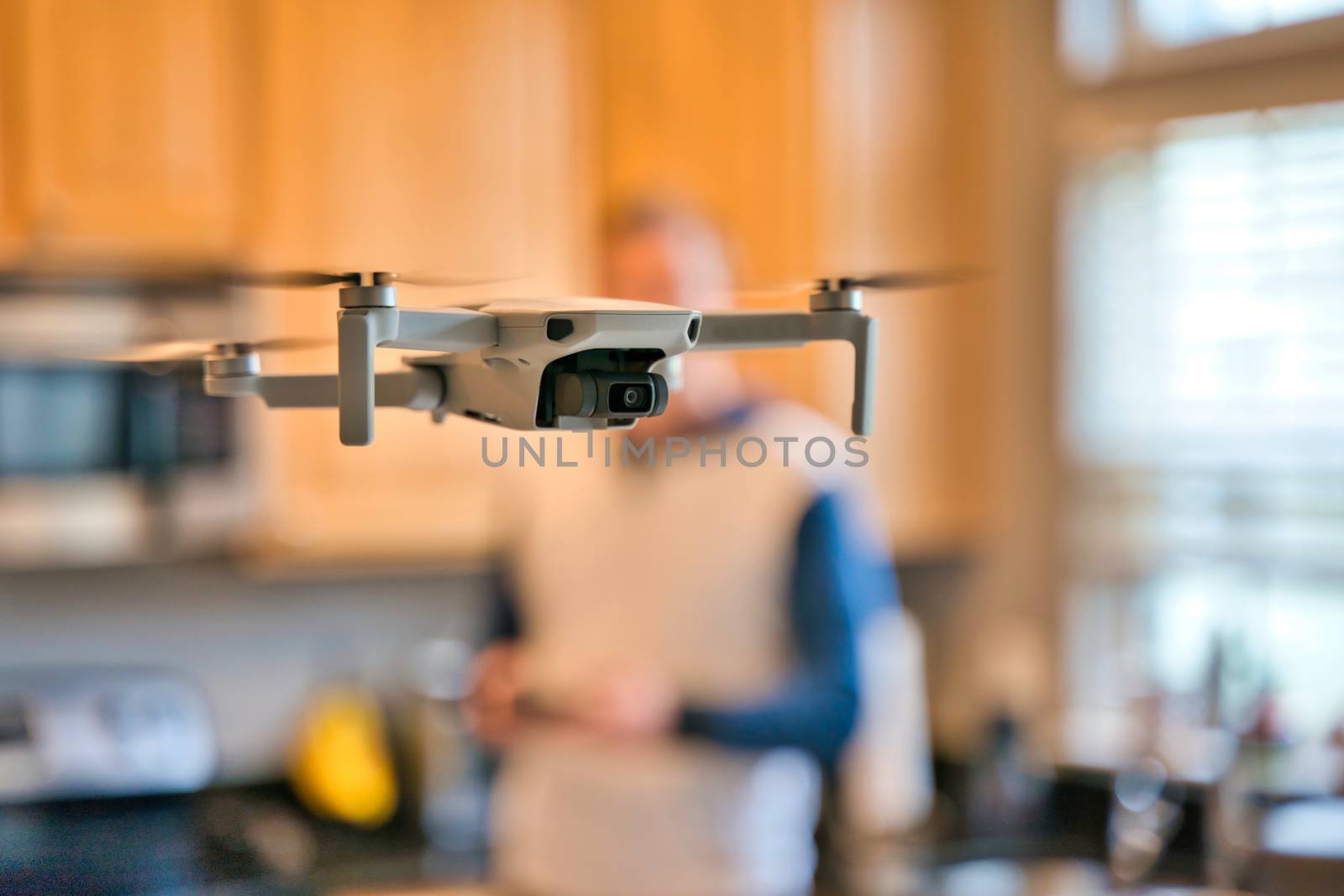 Drone flying indoors with kitchen and pilot visible in background. Amateur drone flying inside for practice. Pilot with remote visible.