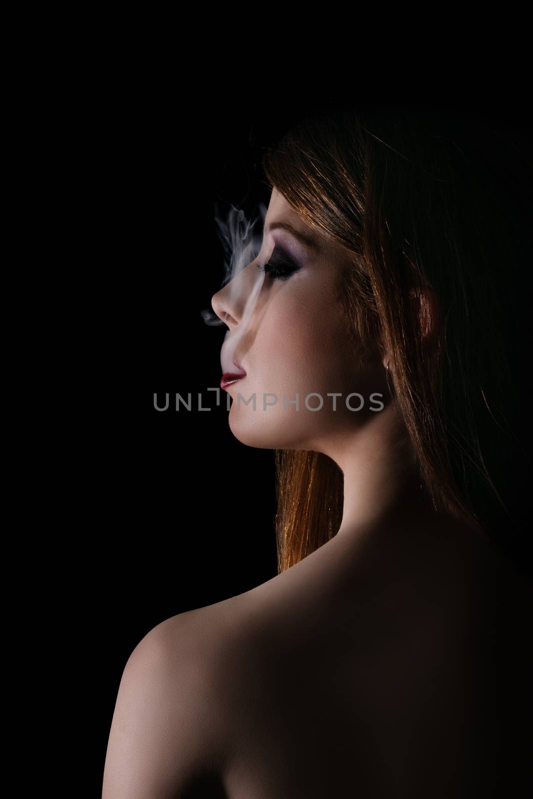 Low key profile shot of a gorgeous young woman with smokey eye makeup and red lipstick, smoke coming out of her mouth, isolated on black background. Smoking concept. Night, beauty, mystery, gothic style, fashion concept.