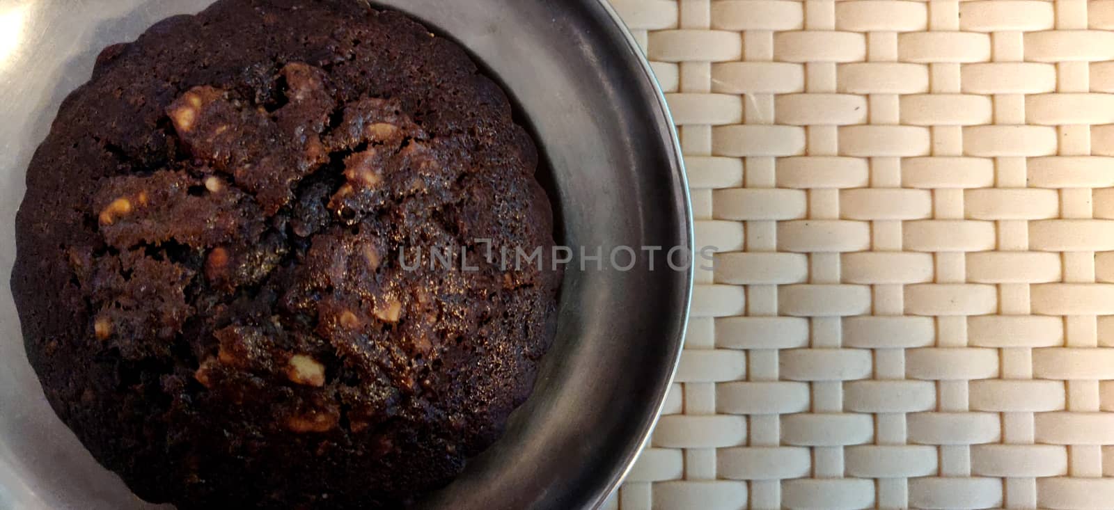 A close up of a big chocolate walnut muffin on a steel plate on a weave pattern table