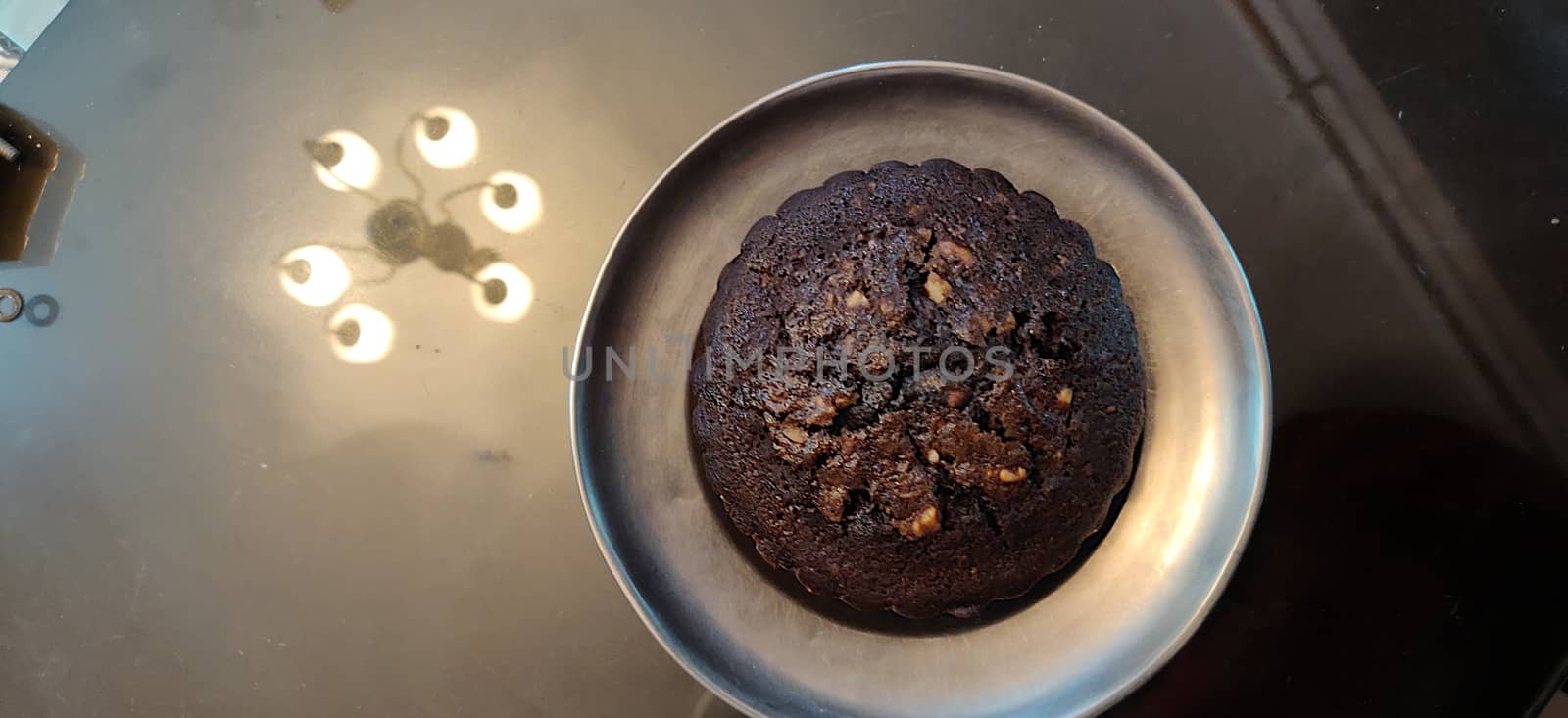 A chocolate walnut muffin on a steel plate against a reflective surface reflecting chandelier lights