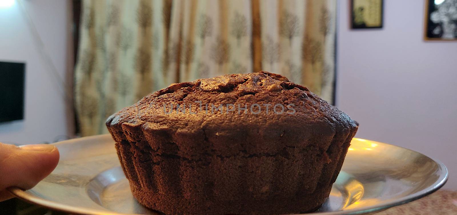A close up of chocolate walnut muffin on a steel plate held up in the air by a hand against the wall of the house