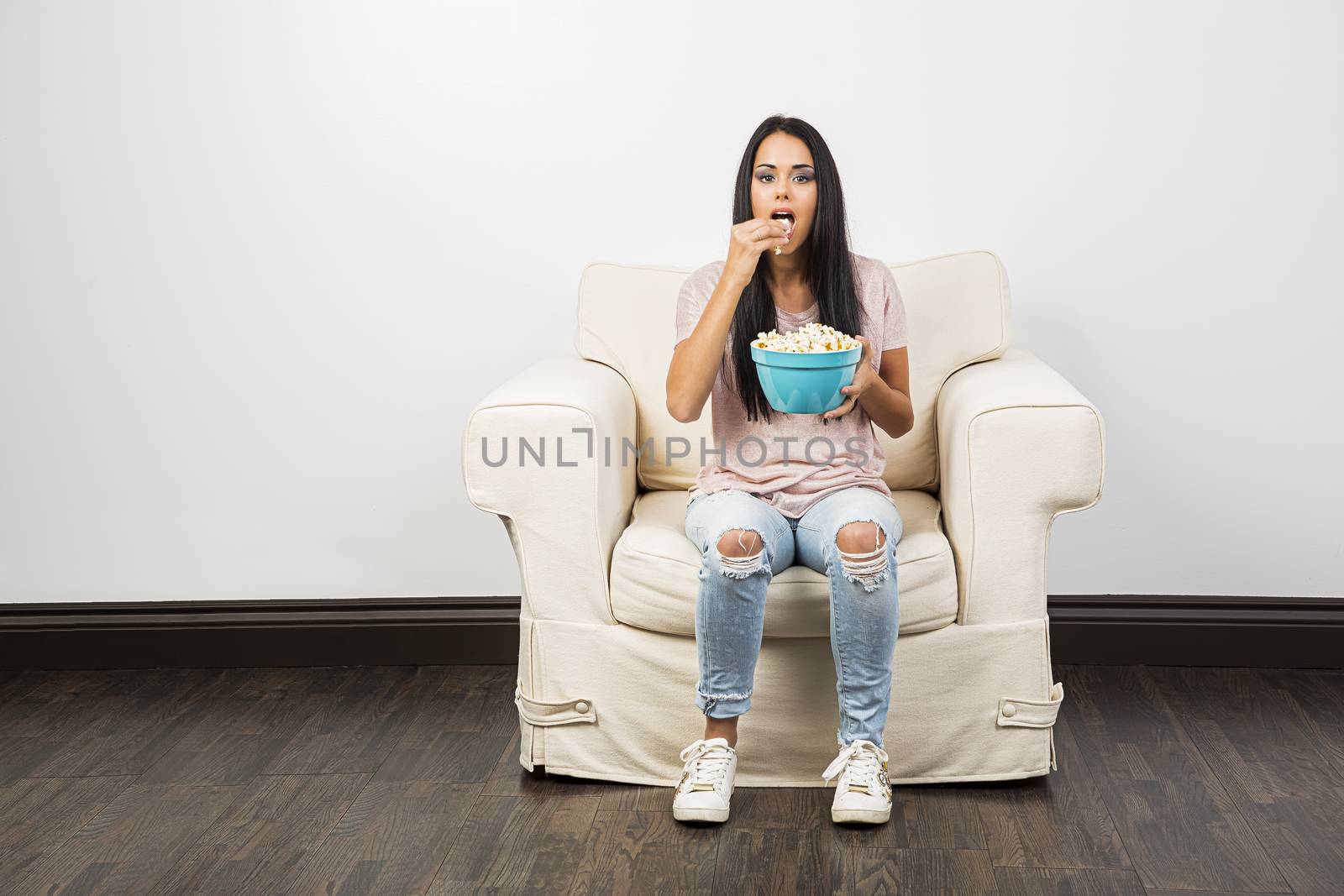 Young woman sitting on a white couch, eating popcorn from a blue bowl