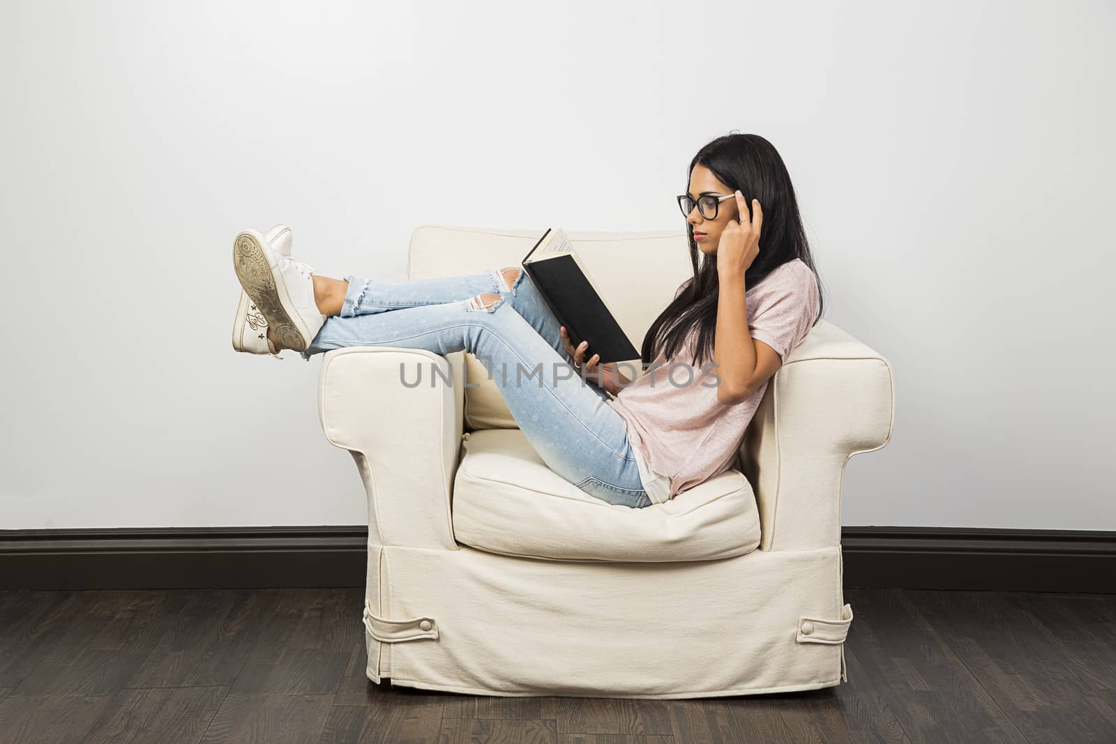 twenty something woman, sitting on a white couch, reading a hard cover book and adjusting her glasses