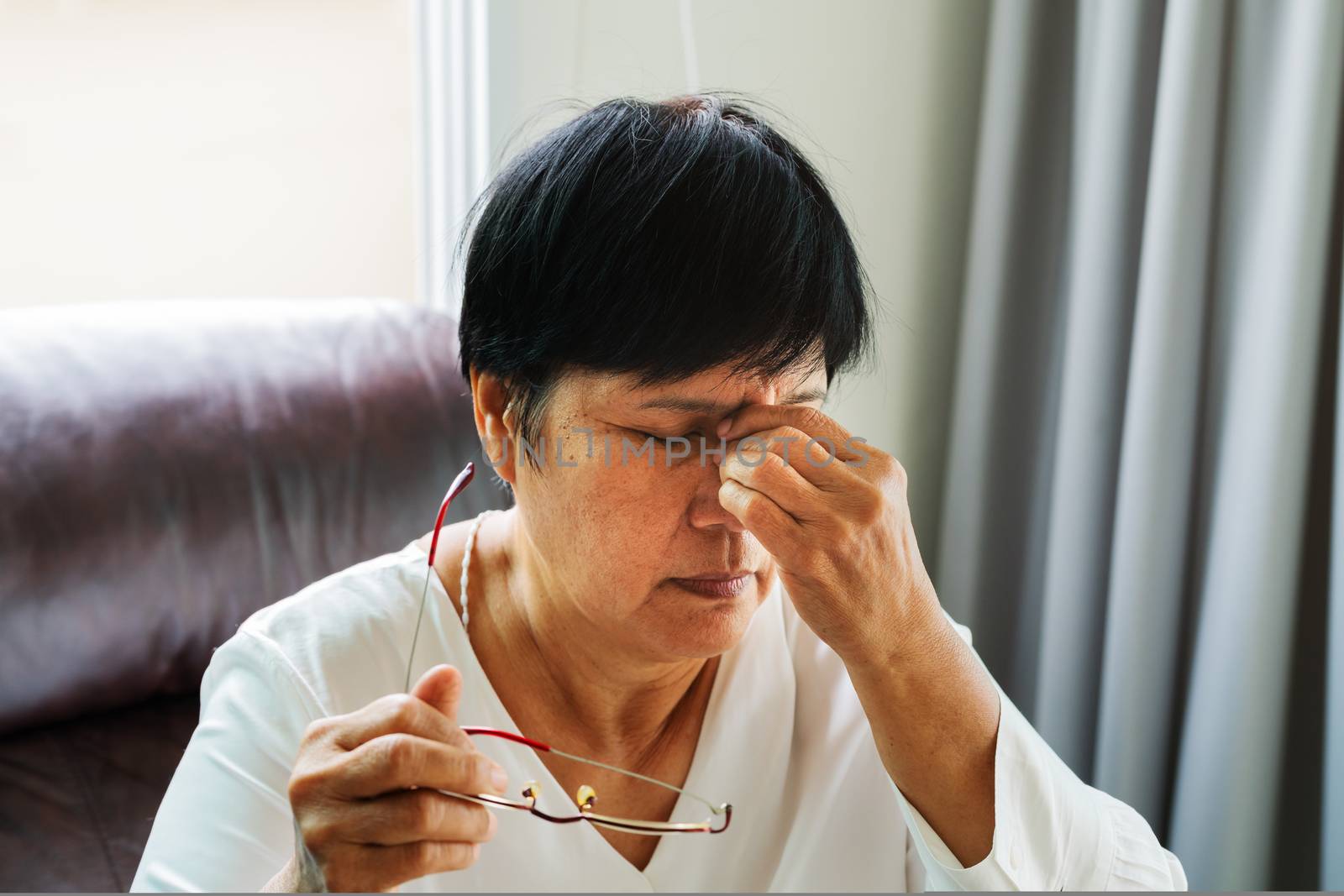 Tired old woman removing eyeglasses, massaging eyes after reading paper book. feeling discomfort because of long wearing glasses, suffering from eyes pain or headache