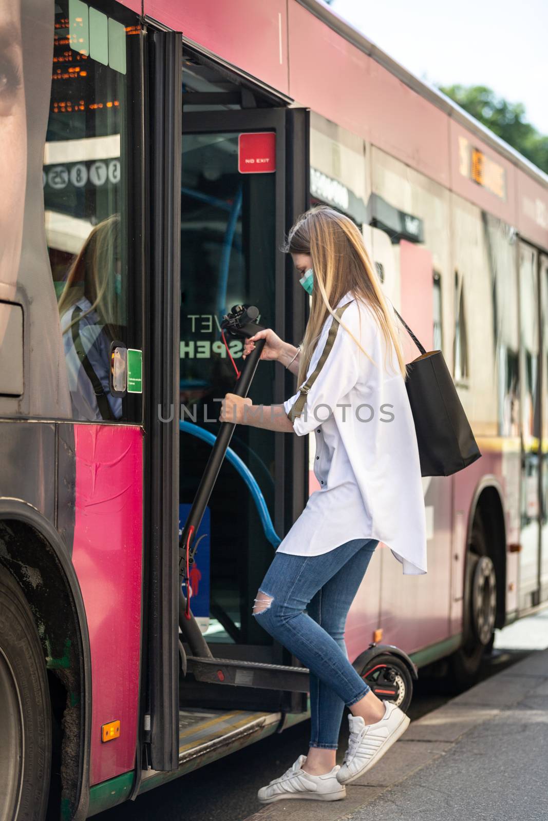 Teenage girl commuter entering city bus carring foldable electric urban scooter, wearing protective face mask against spreading of coronavirus and disease transmission at time of covid-19 pandemic.