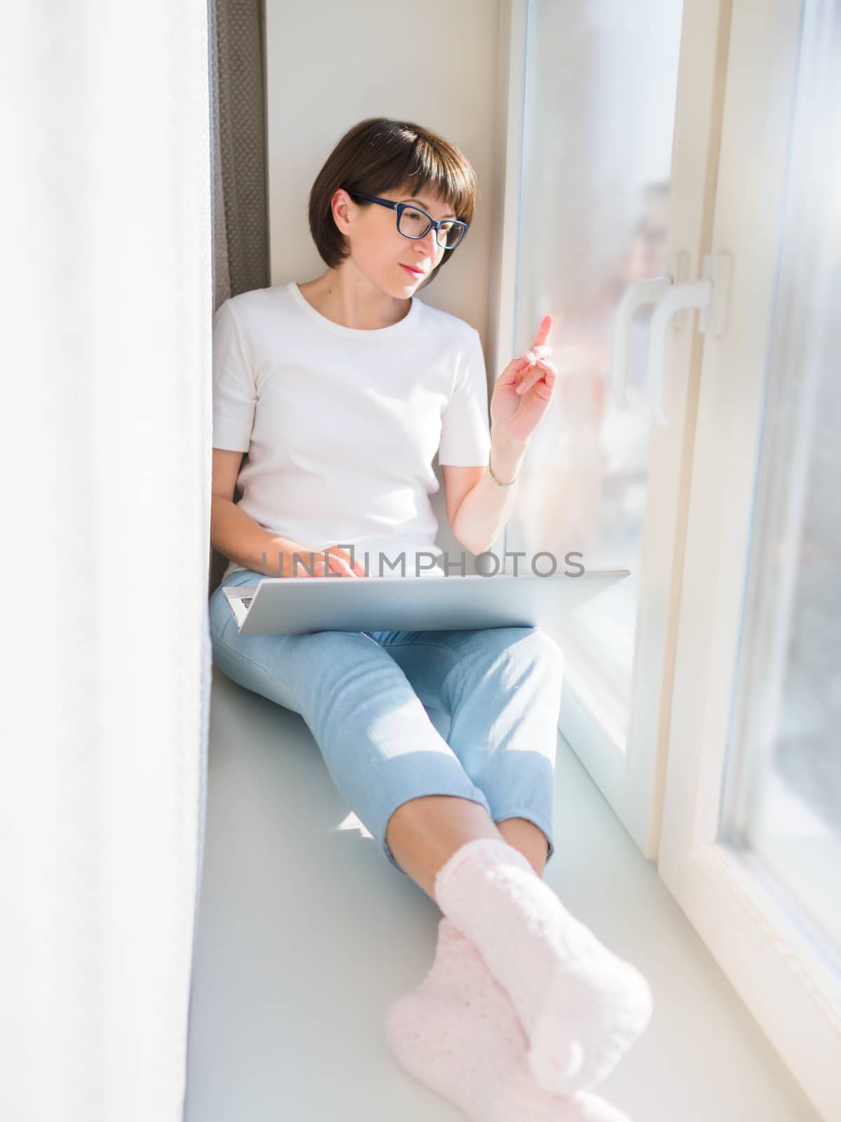 Woman works remotely from home. She sits on window sill with lap by aksenovko